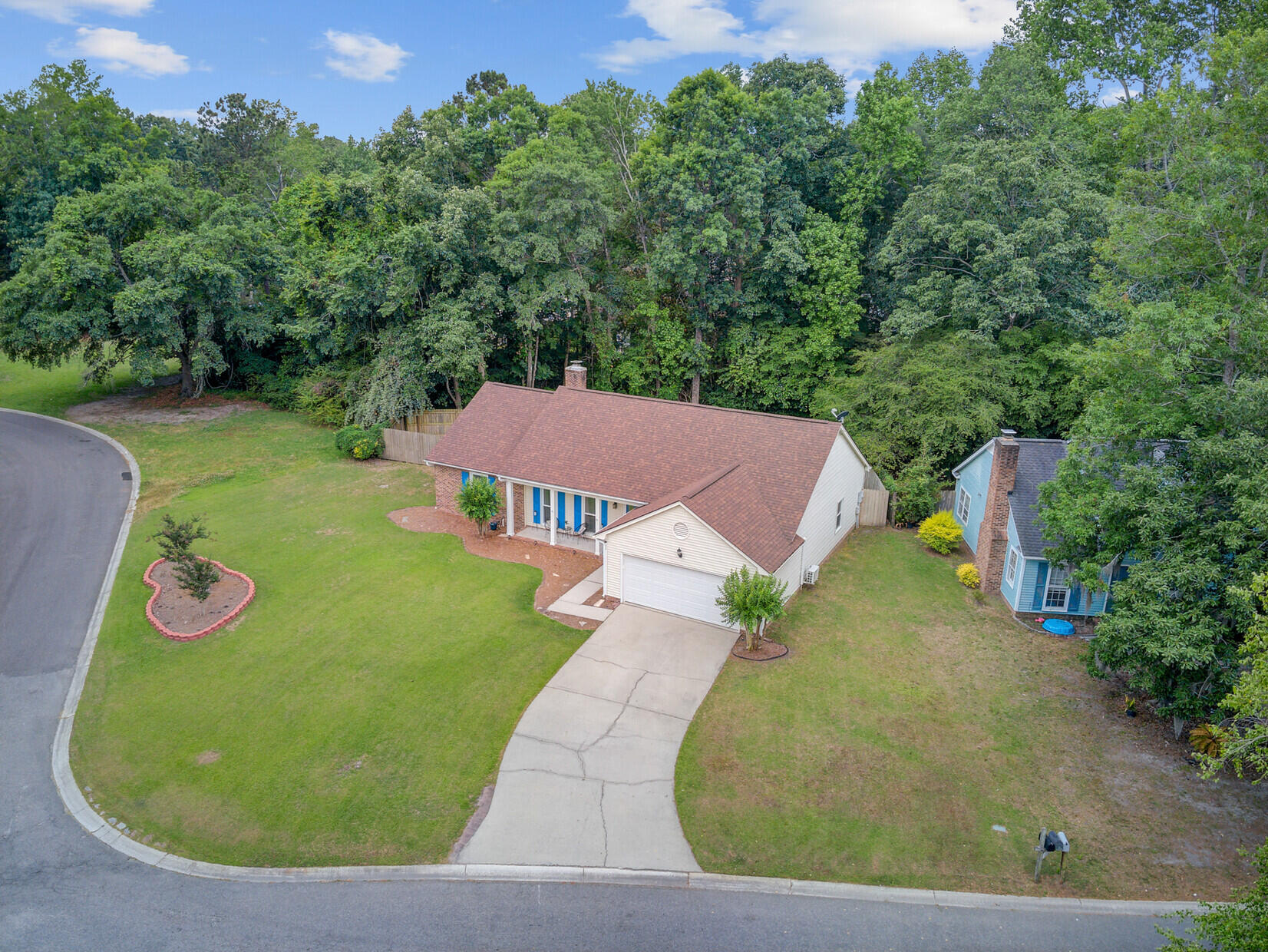 Aerial view of a single-story house with a curved driveway, broad lawn, and wooded backdrop.