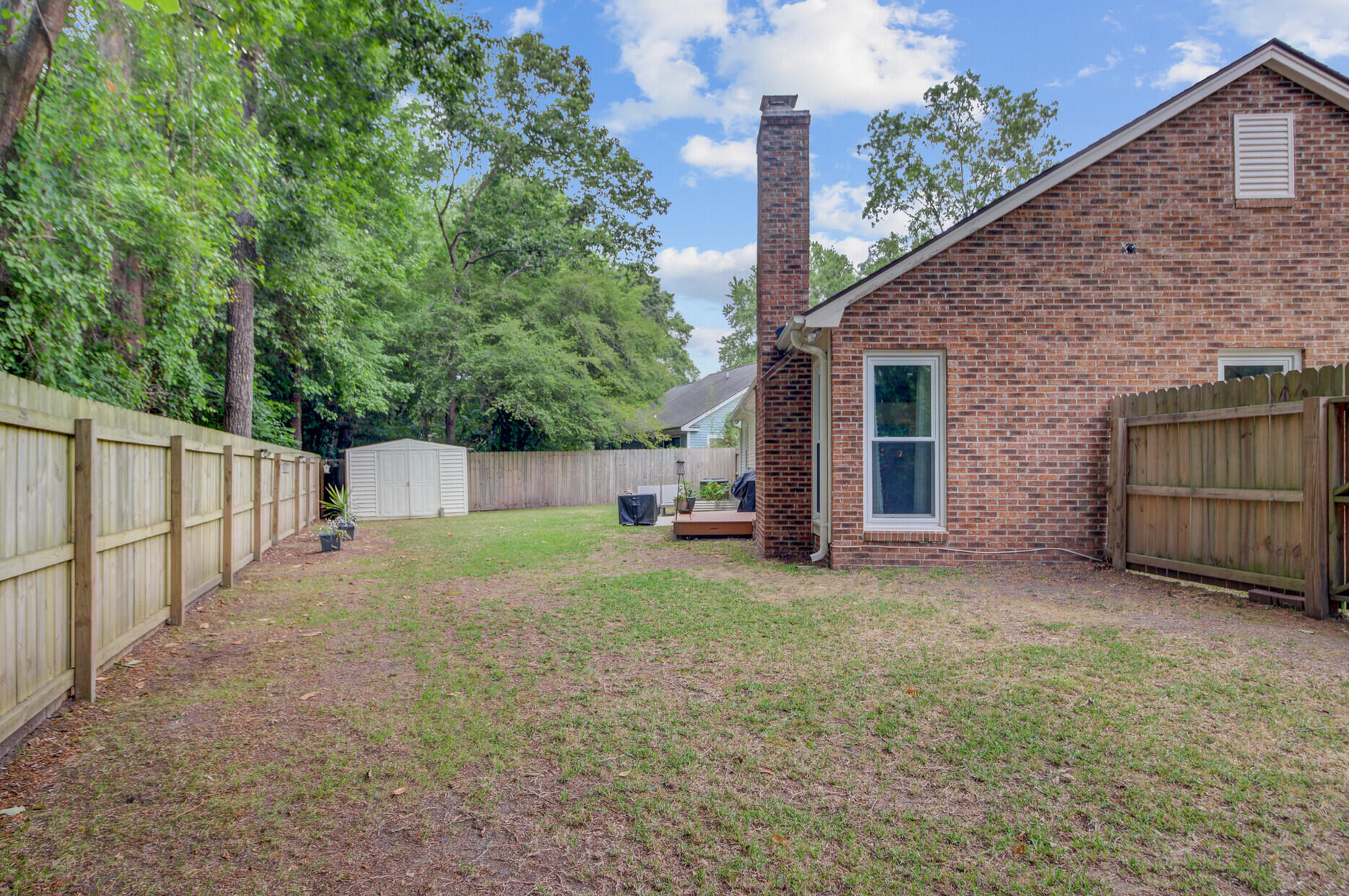Fenced backyard beside a red brick house, with a storage shed, small deck, and mature trees.
