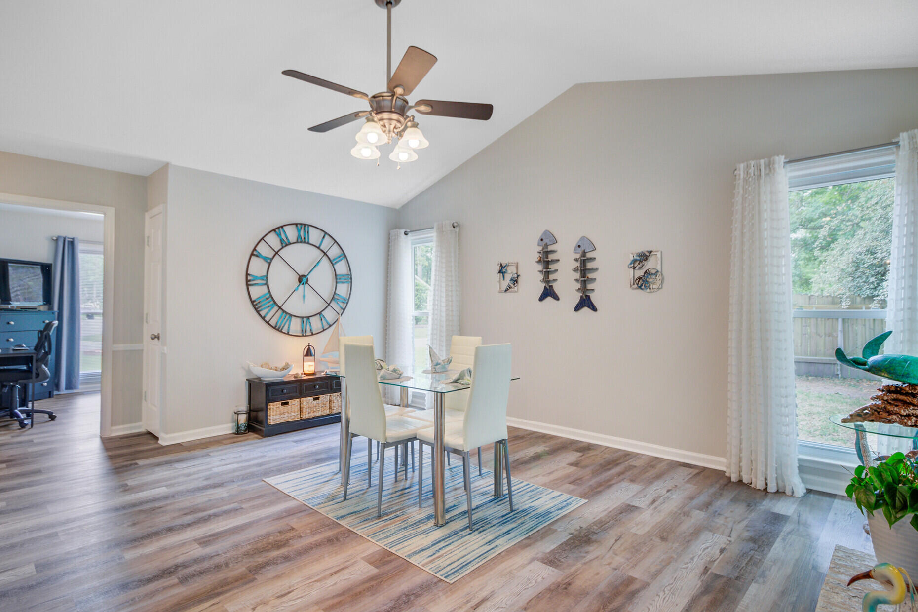 Dining room with a glass table, white chairs, oversized clock, and large curtained windows.
