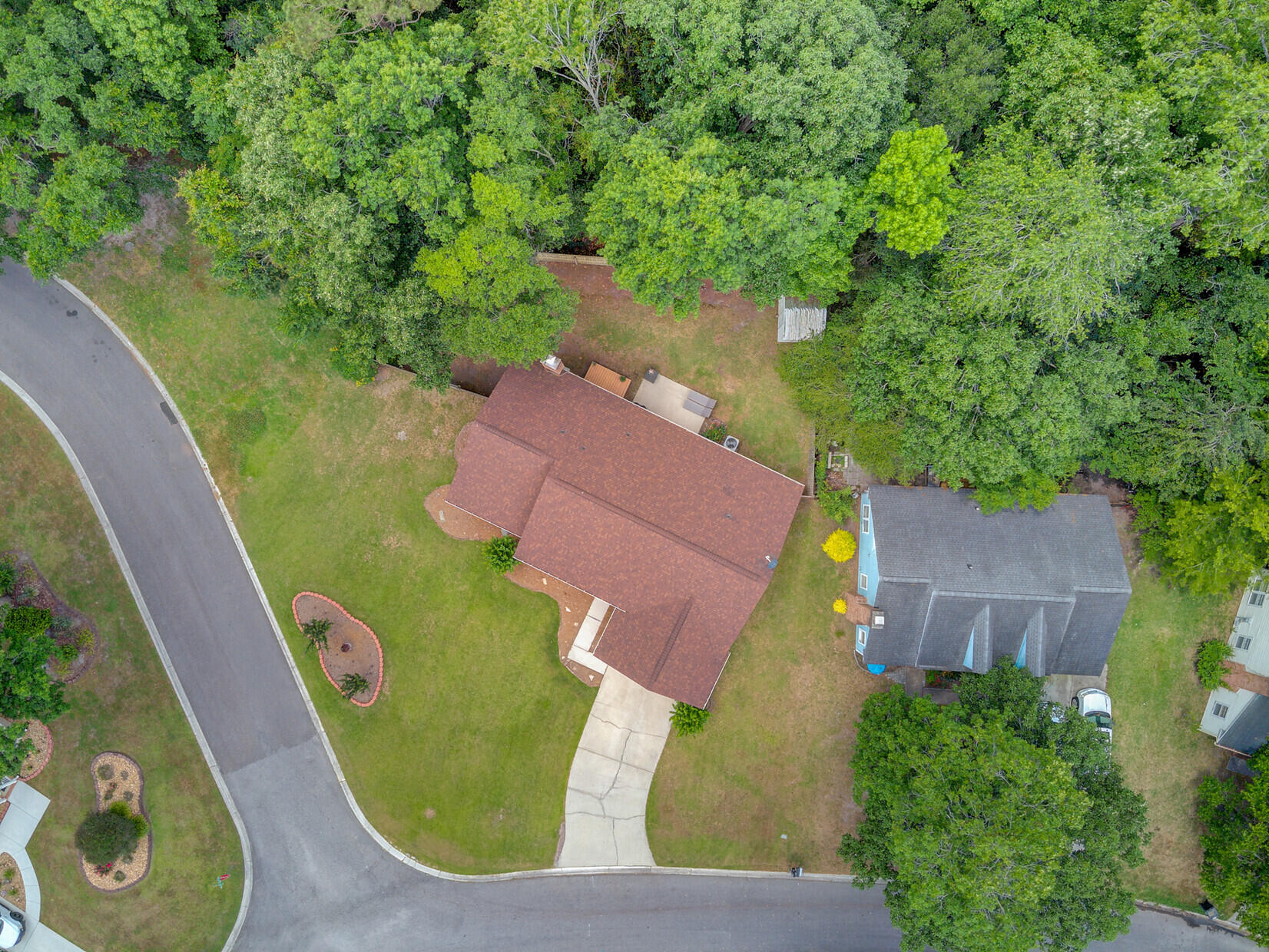 Aerial view of a home with a reddish-brown roof, driveway, lawn, and mature trees.
