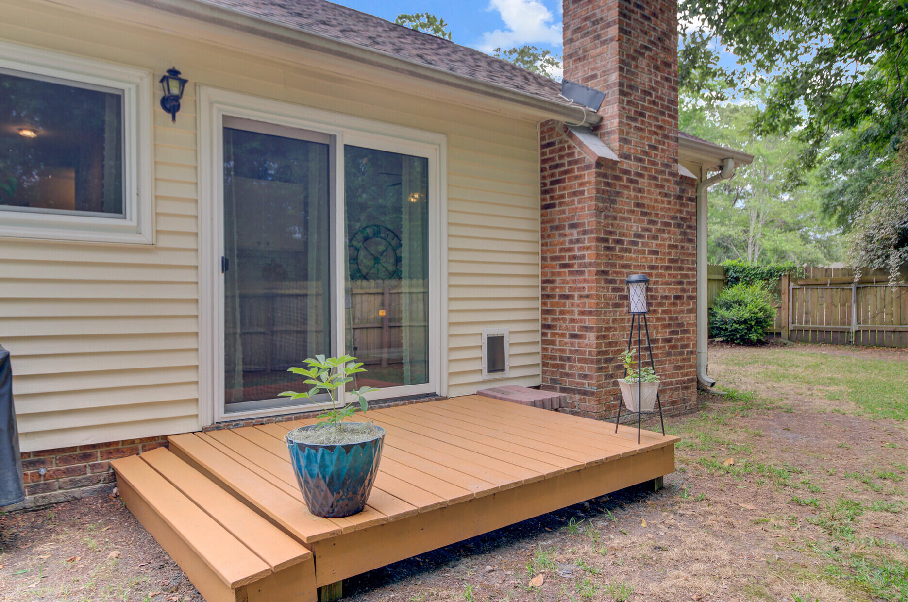 Raised wooden deck beside sliding glass doors and a tall brick chimney, overlooking a fenced yard.
