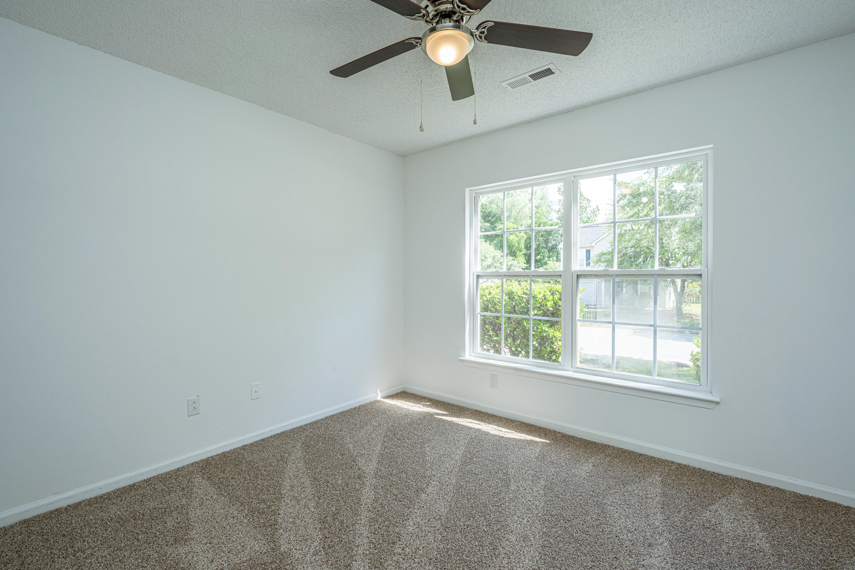 Empty carpeted bedroom with a ceiling fan and a large window overlooking greenery.