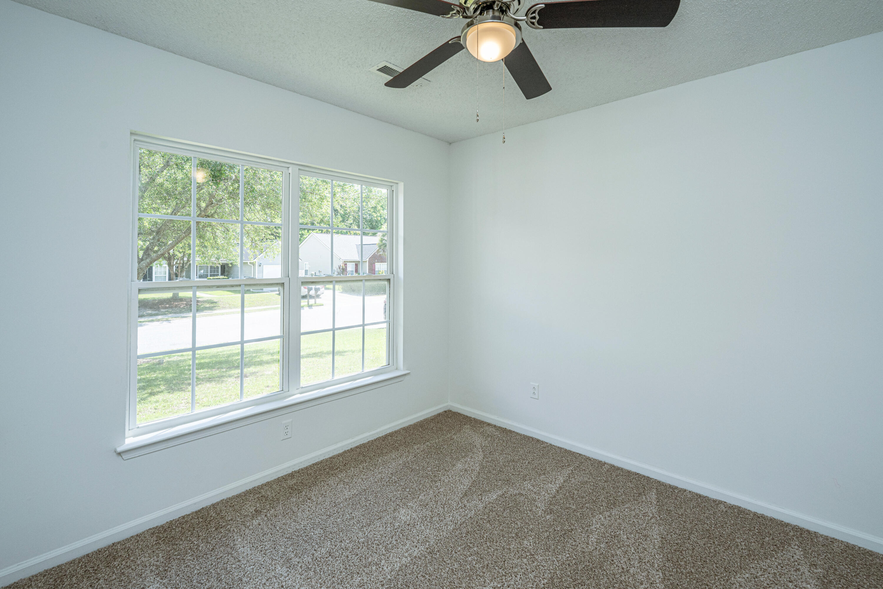Empty bedroom with beige carpet, ceiling fan, and large windows overlooking a leafy street.