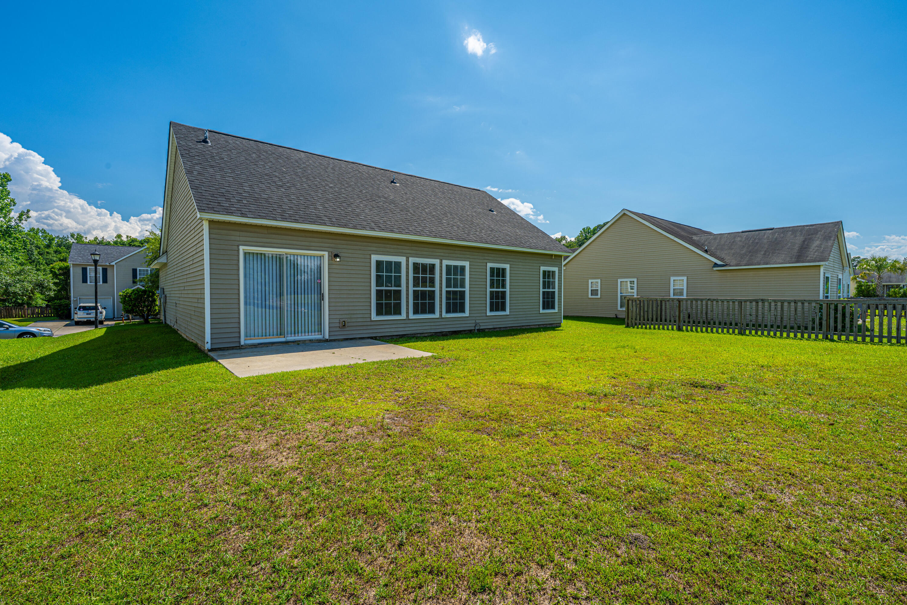 Rear of a gray-sided house with a sliding glass door, concrete patio, and broad sunny lawn.