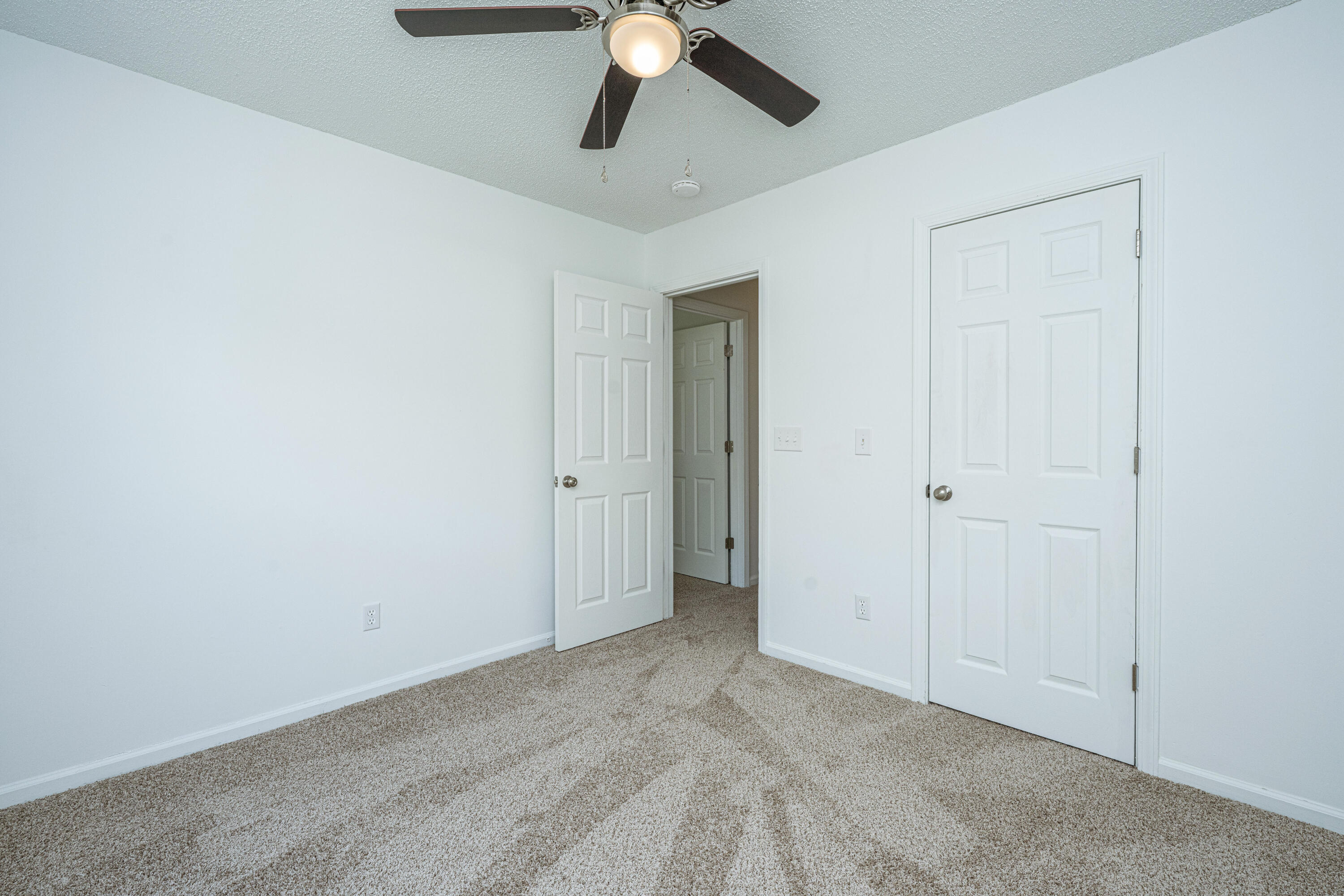 Empty bedroom with beige carpet, a ceiling fan, and an open door to the hallway.