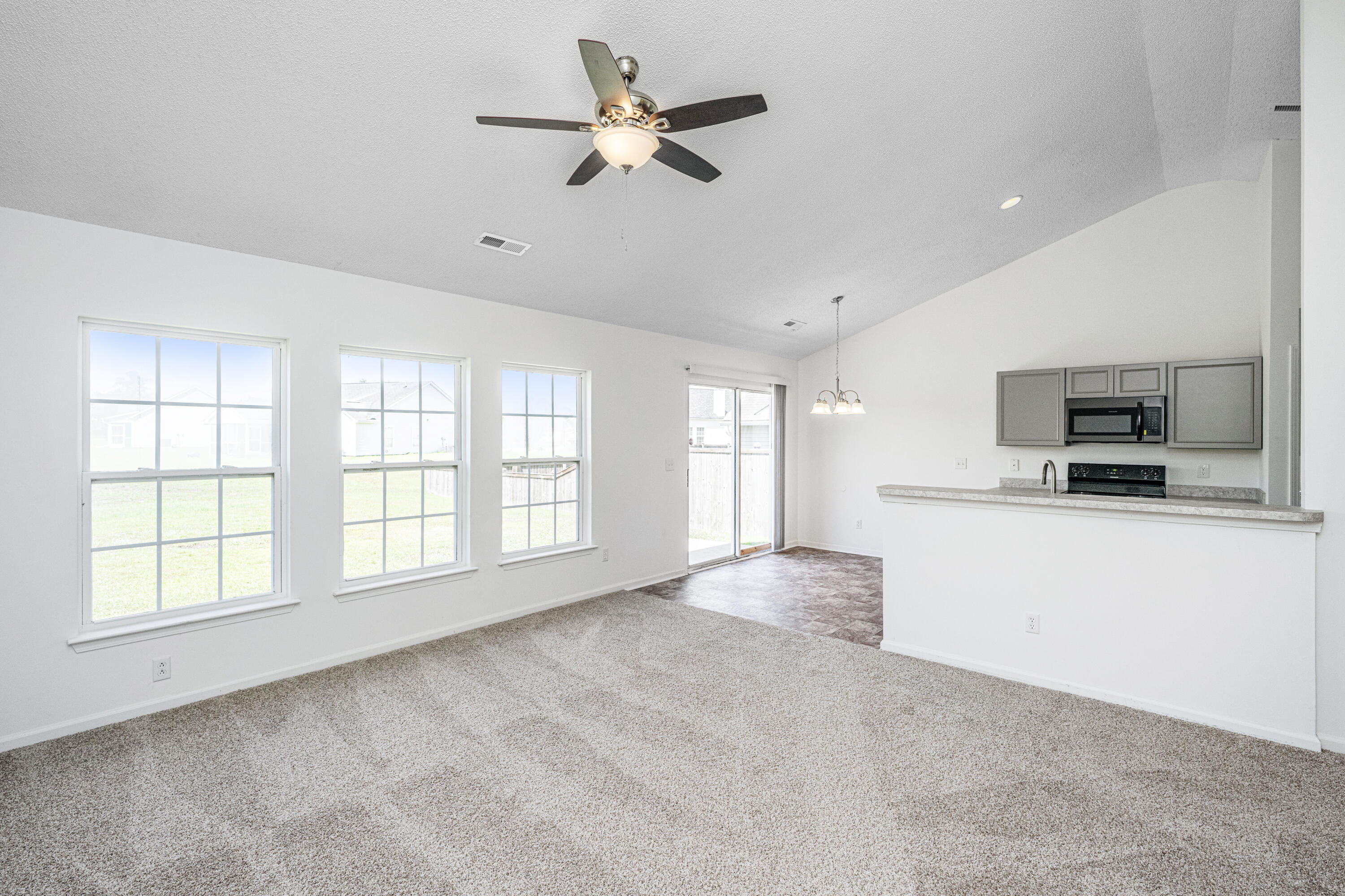 Empty living area with three large windows, ceiling fan, and an open kitchen.