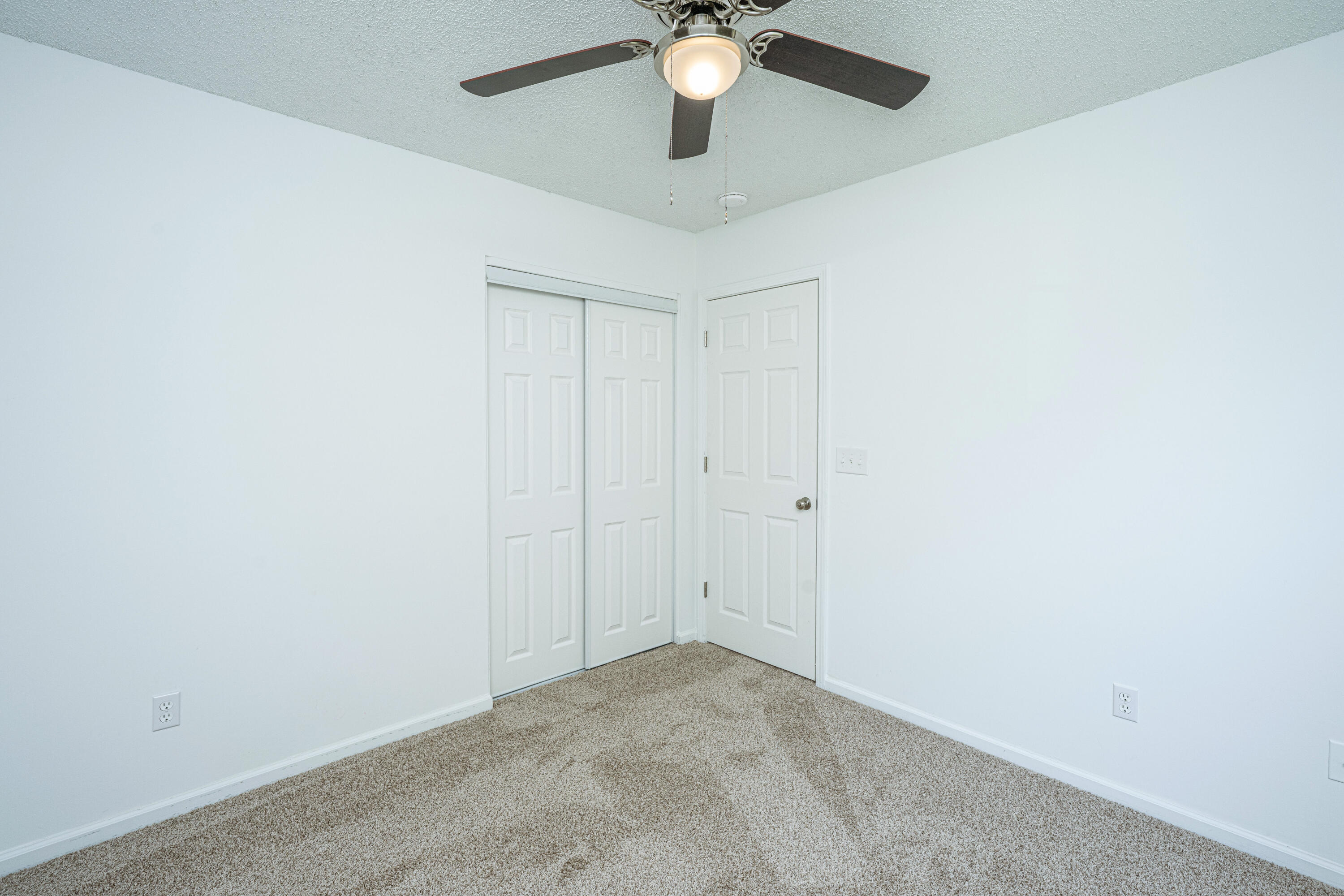 Empty bedroom with beige carpet, white sliding closet doors, and a ceiling fan with light.