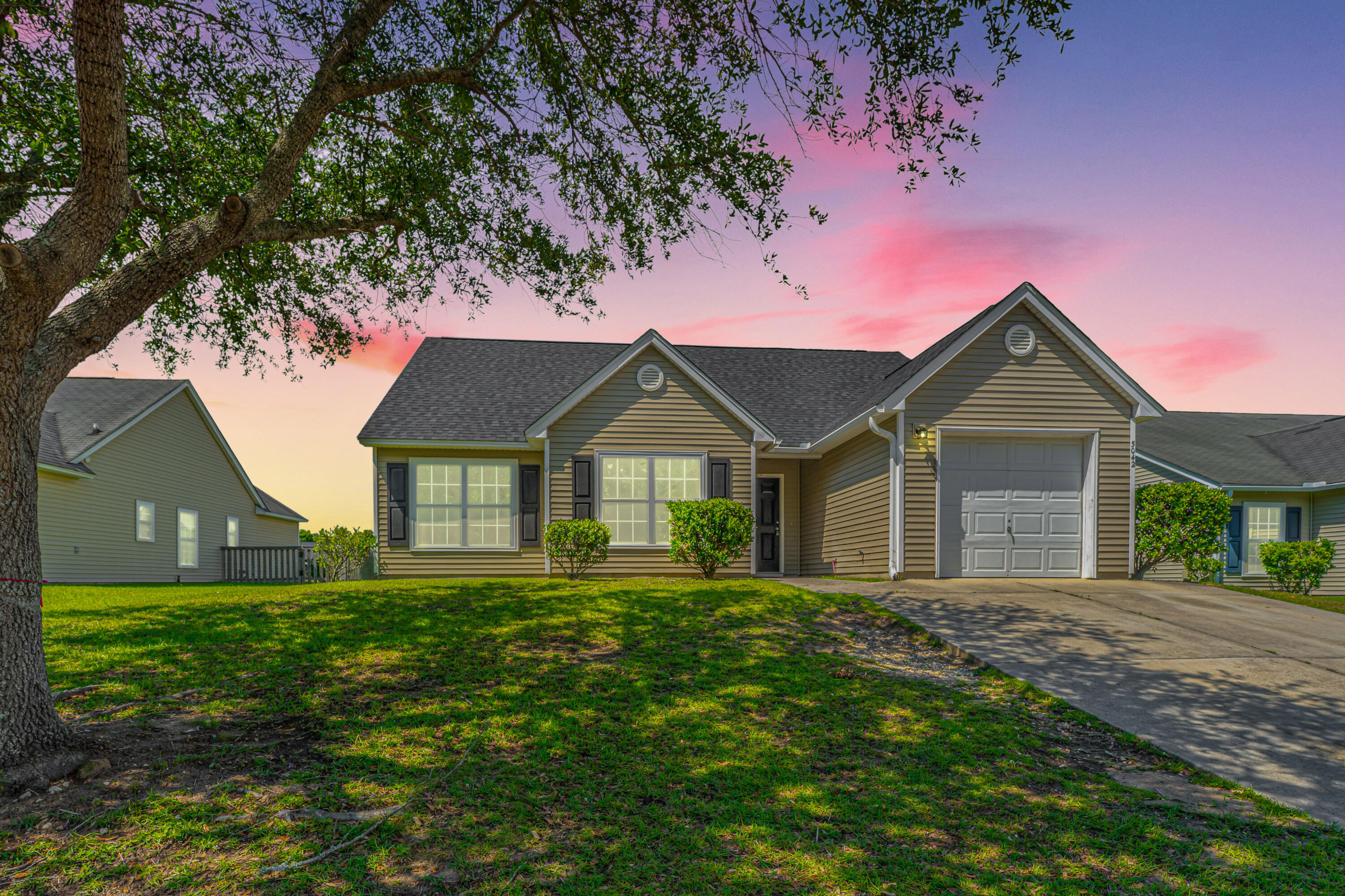 Single-story olive-gray house with an attached garage, broad lawn, and driveway beneath a pink dusk sky.