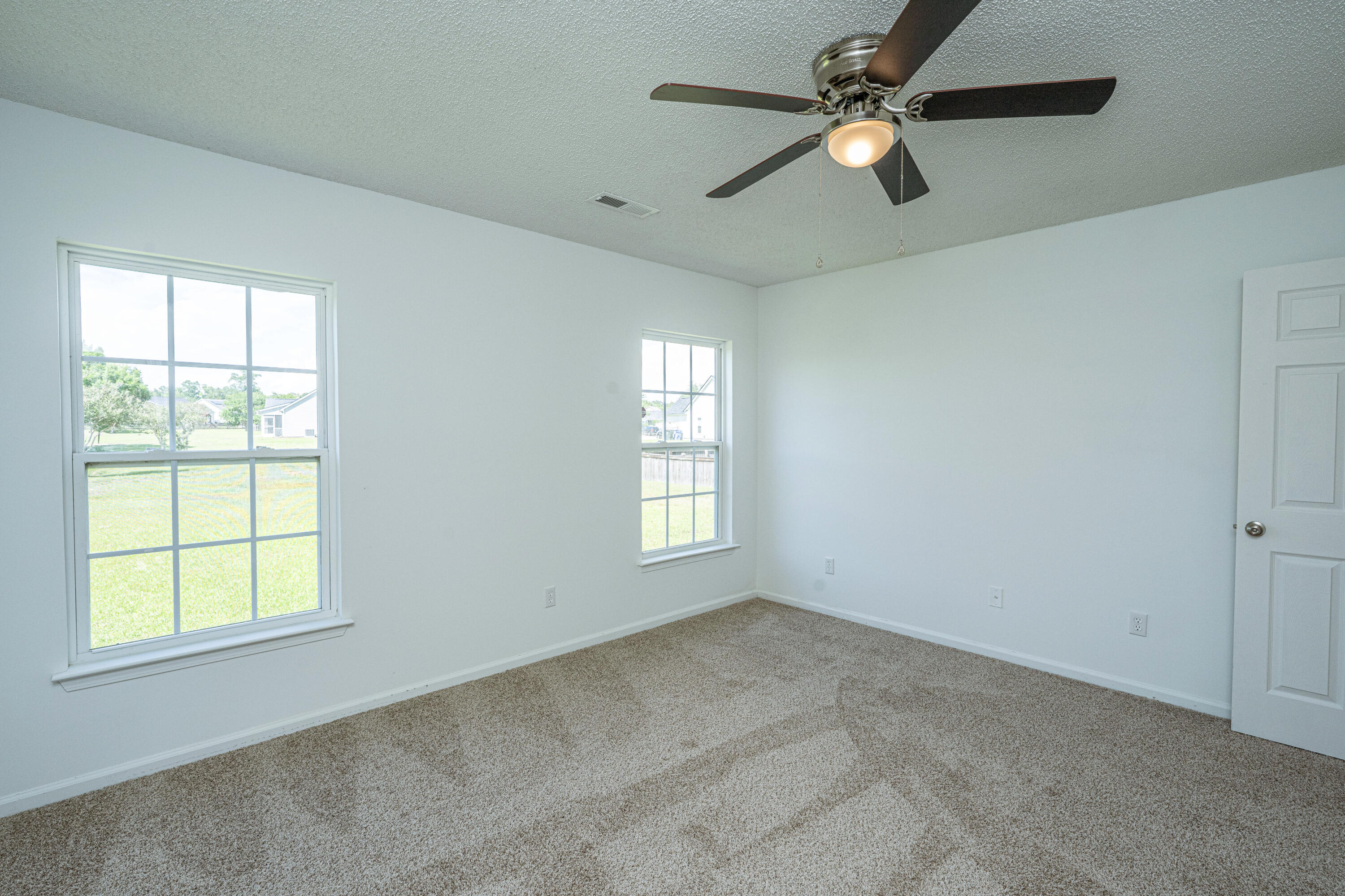Empty bedroom with beige carpet, two yard-facing windows, and a ceiling fan with light.