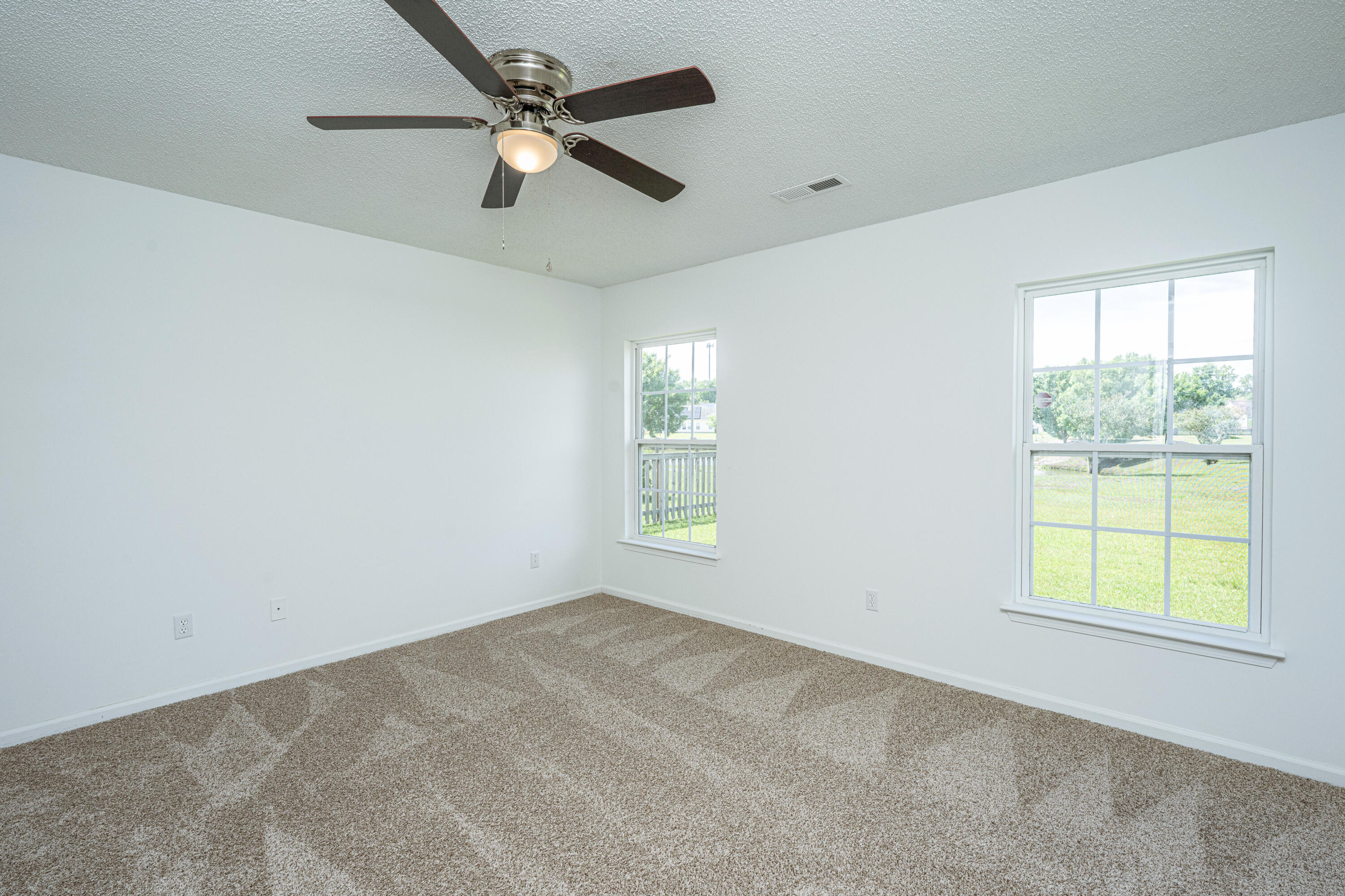 Empty carpeted bedroom with a ceiling fan and two windows overlooking a lawn.