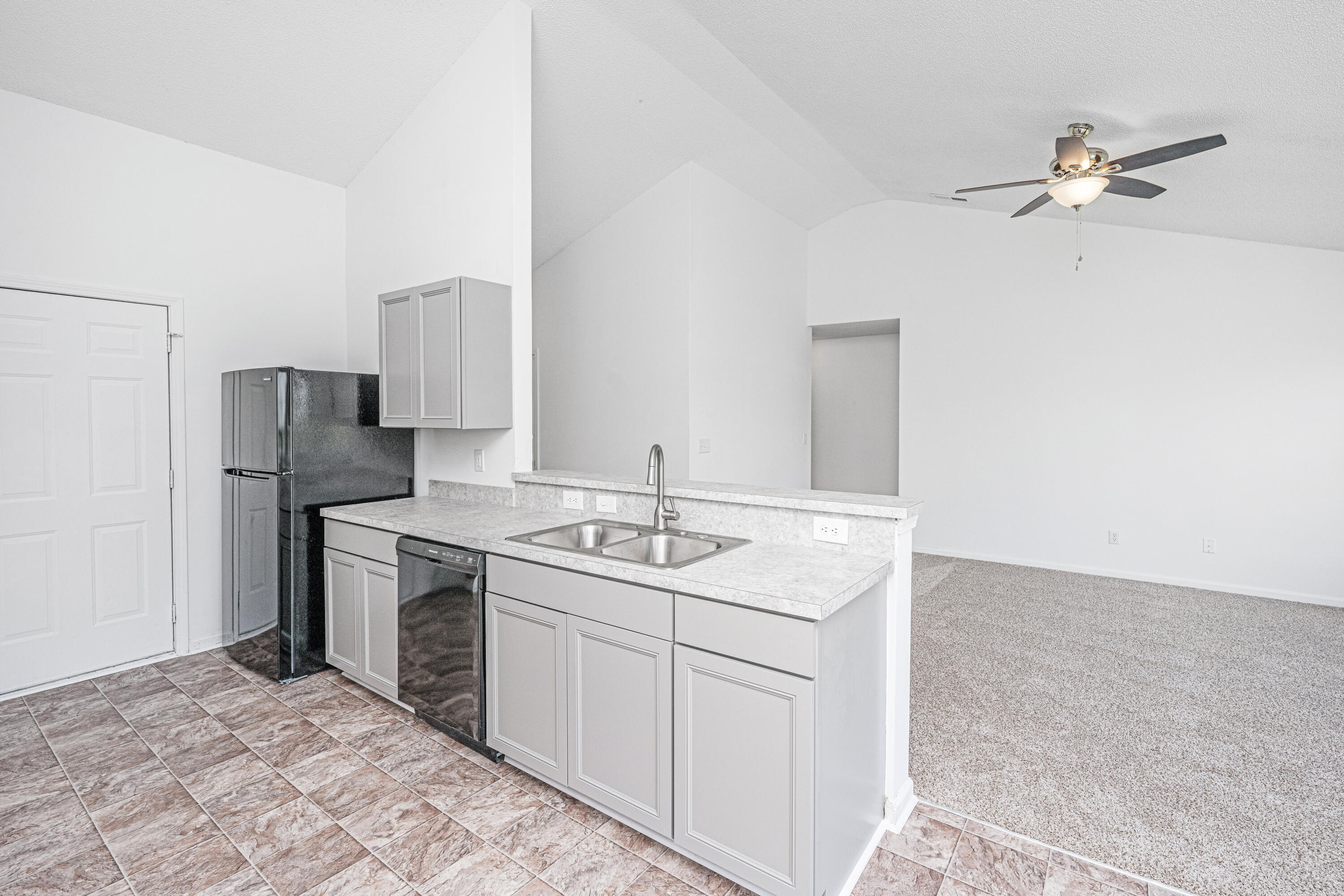 Empty kitchen with gray cabinets, a double-basin sink, and black appliances opening to a carpeted living area.