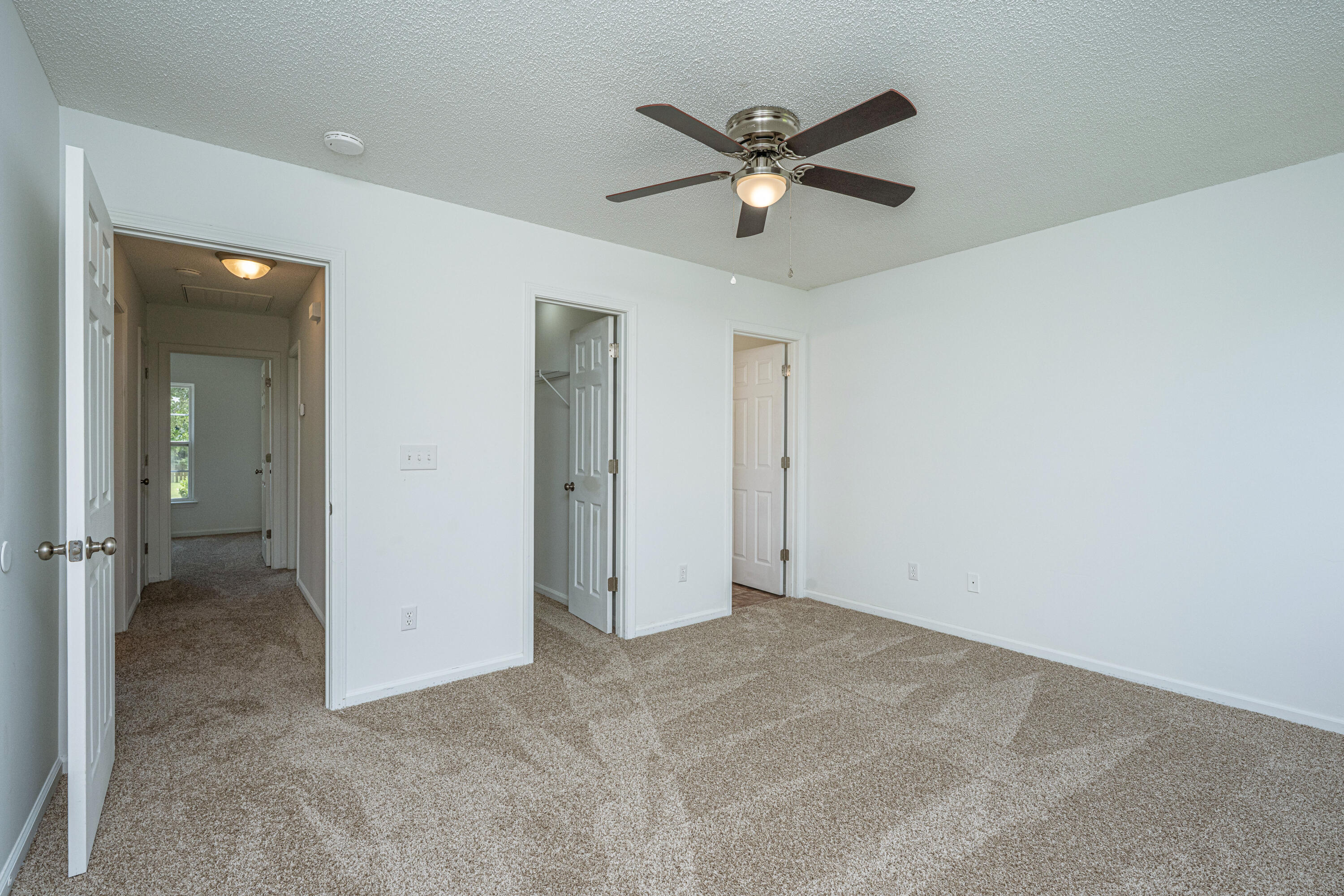 Empty carpeted bedroom with a ceiling fan and open doors to the hallway and closets.