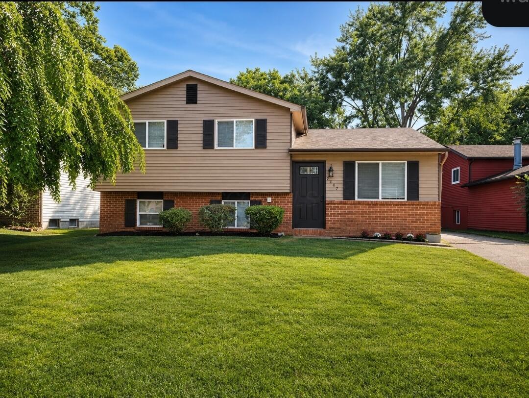 Split-level house with tan siding, brick lower facade, dark shutters, and a broad green lawn.