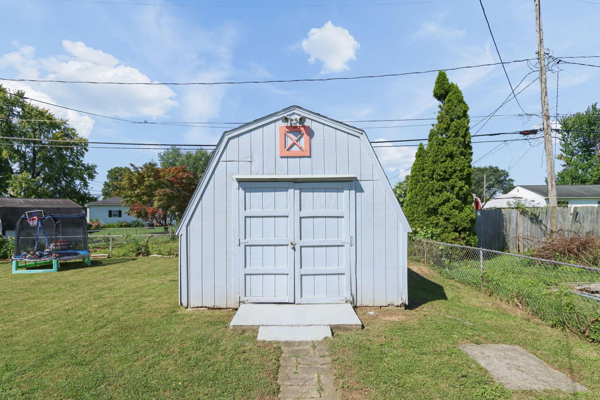 White gambrel-roof shed with double doors in a grassy backyard beside a trampoline.