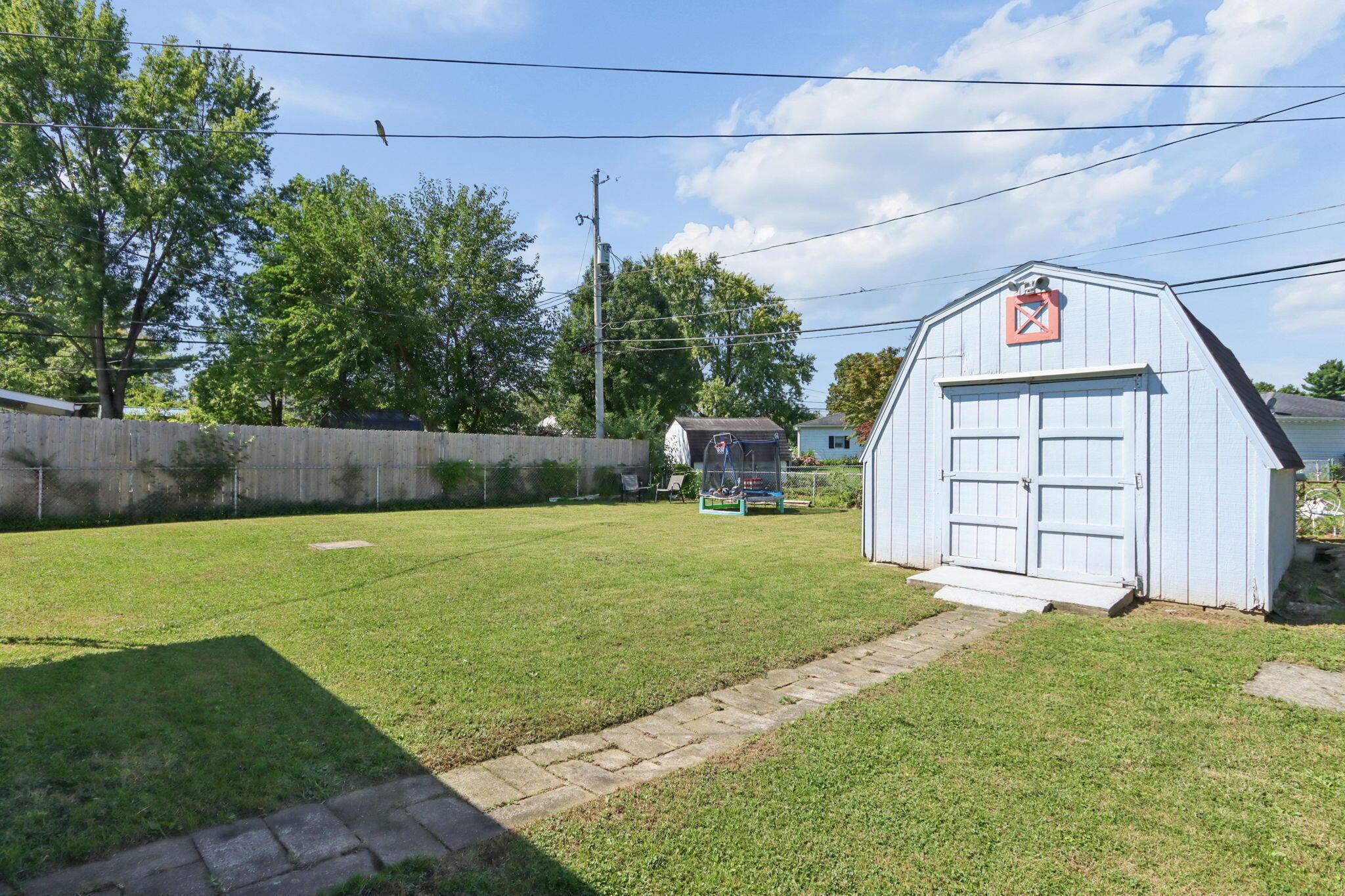 Backyard lawn with a light-blue barn-style shed, paved path, and trampoline.