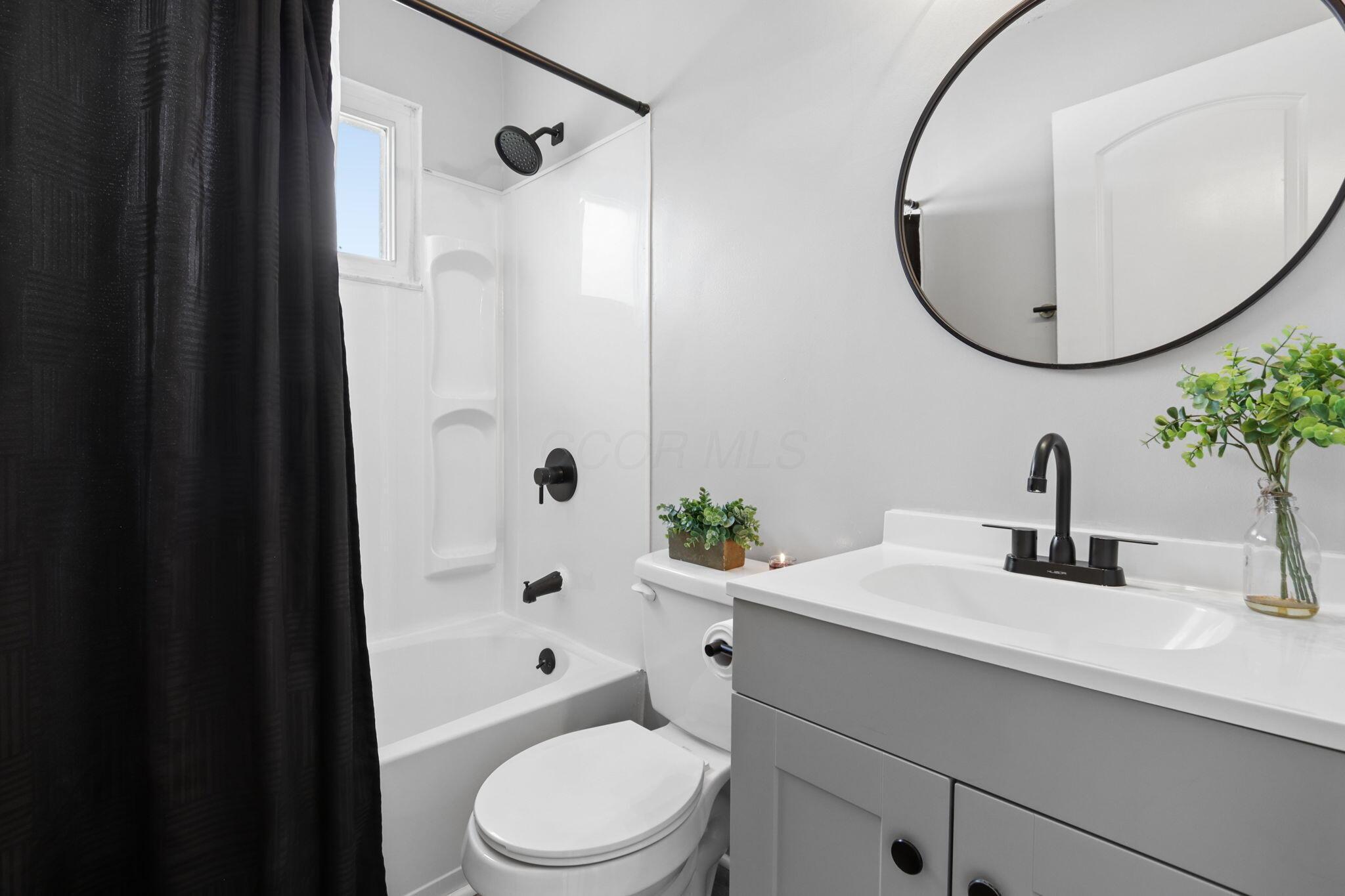 Bathroom with a white tub, black fixtures, gray vanity, and large round mirror.