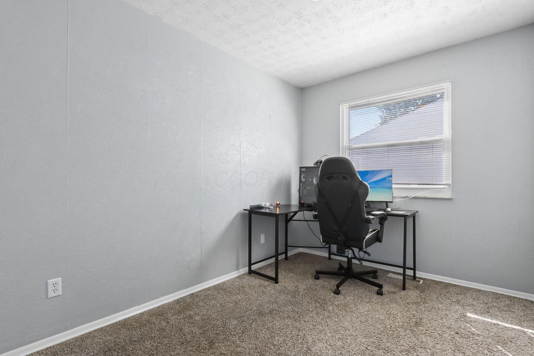 Home office with a black desk, computer monitors, and gaming chair beneath a window with blinds.