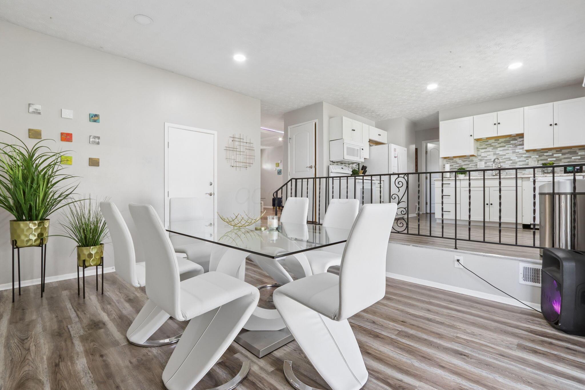 Dining area with a glass-top table and white chairs overlooking a white-cabinet kitchen.