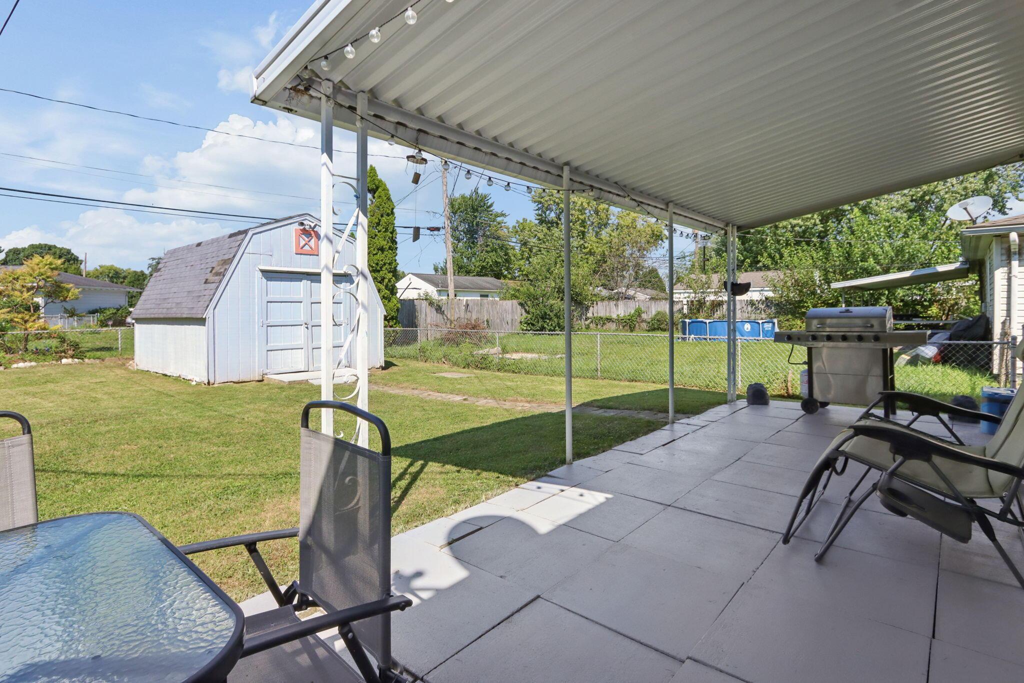 Covered backyard patio with outdoor seating and grill, overlooking a fenced lawn and storage shed.