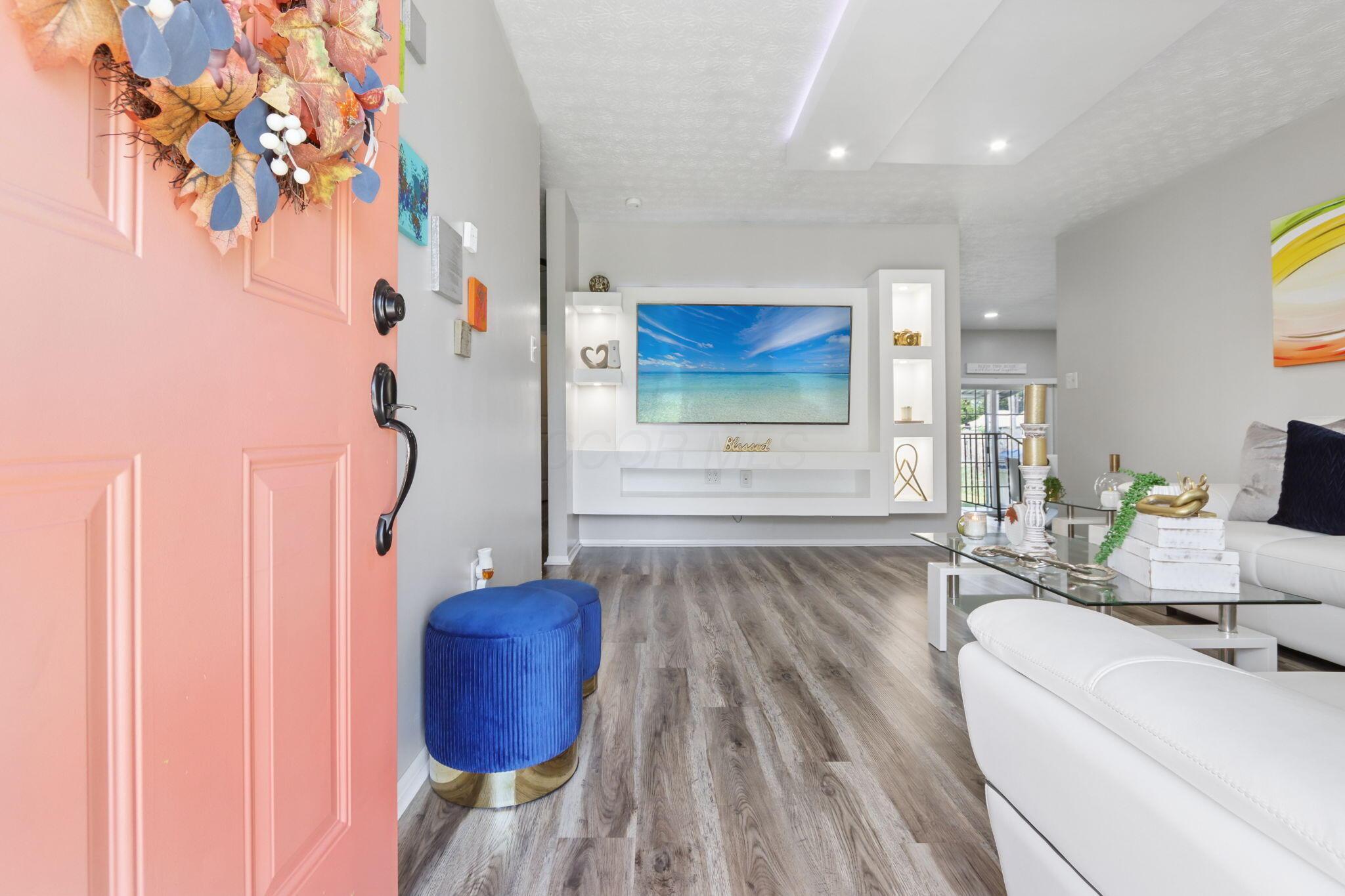 Furnished living room viewed through a coral-pink front door, with a blue ottoman and white entertainment wall.