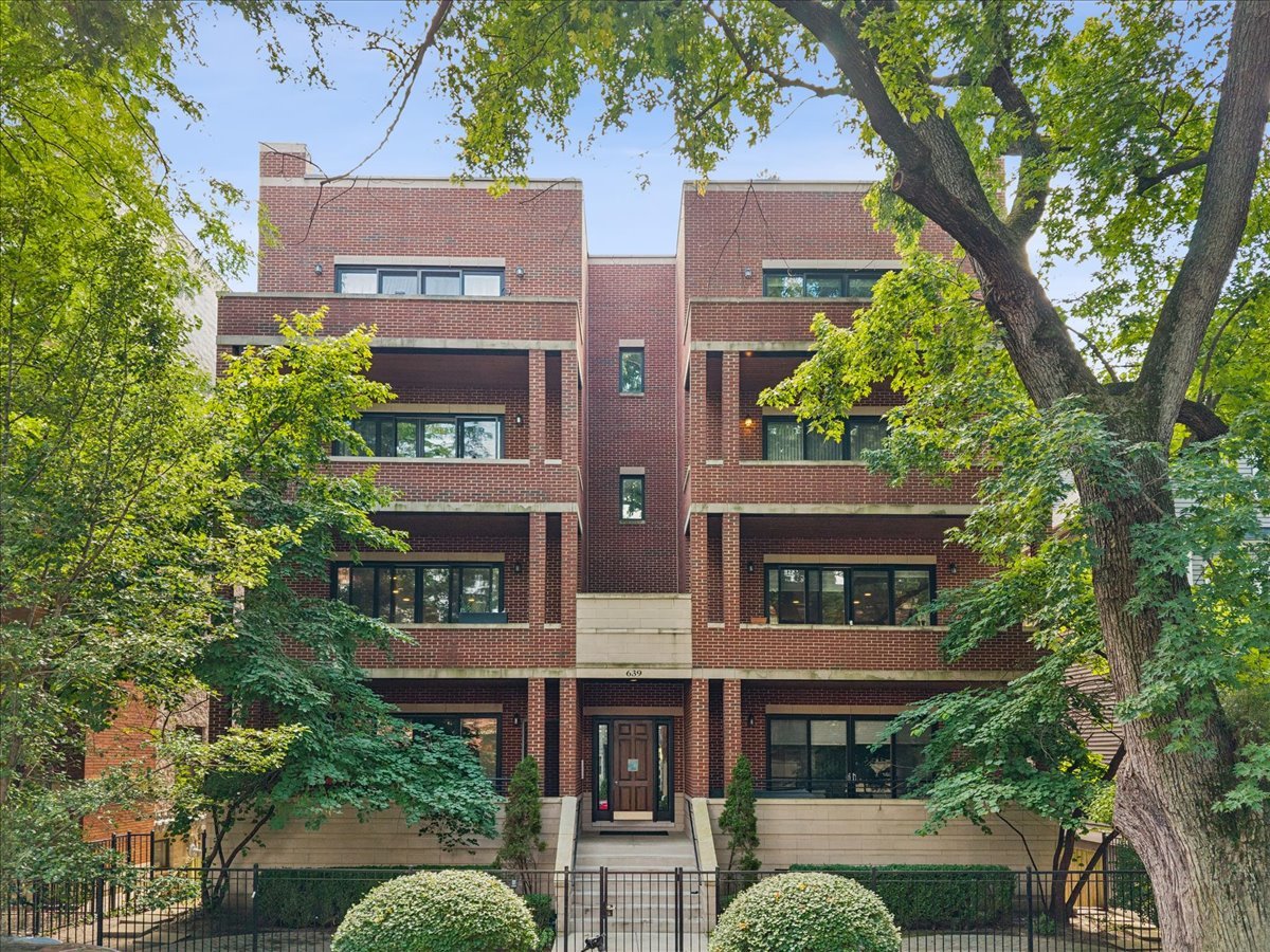 Red brick condominium building with recessed balconies, leafy trees, and a gated entrance.