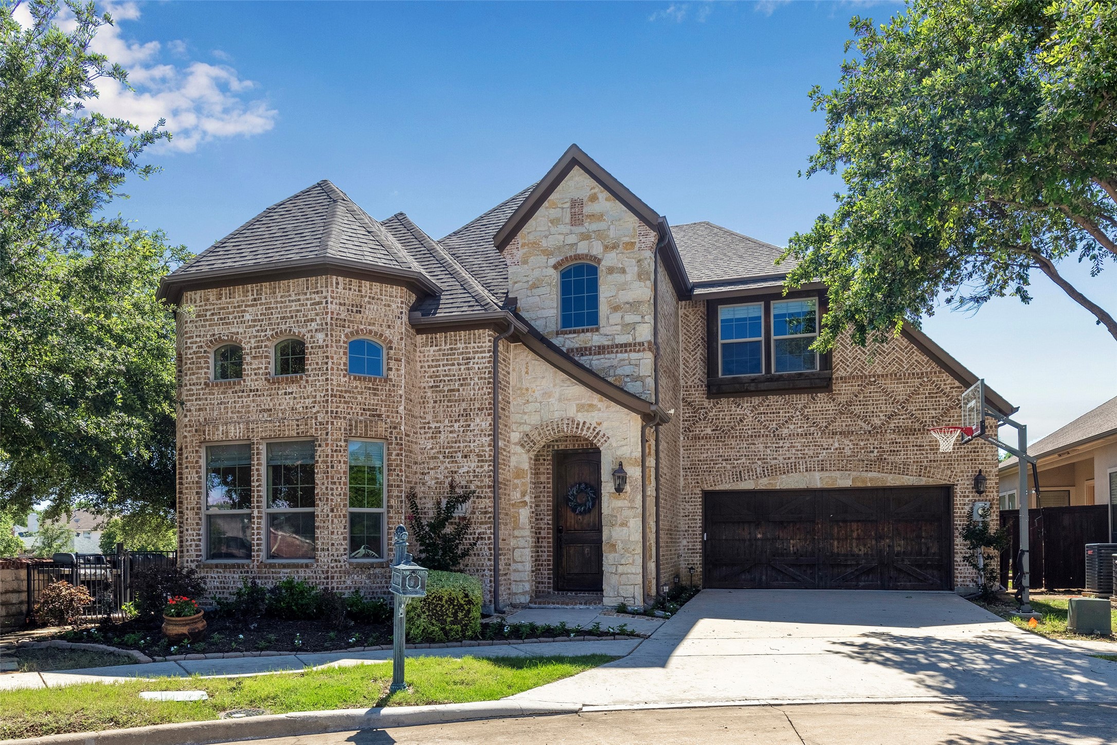 Two-story brick and stone house with a landscaped yard, double garage, and basketball hoop.