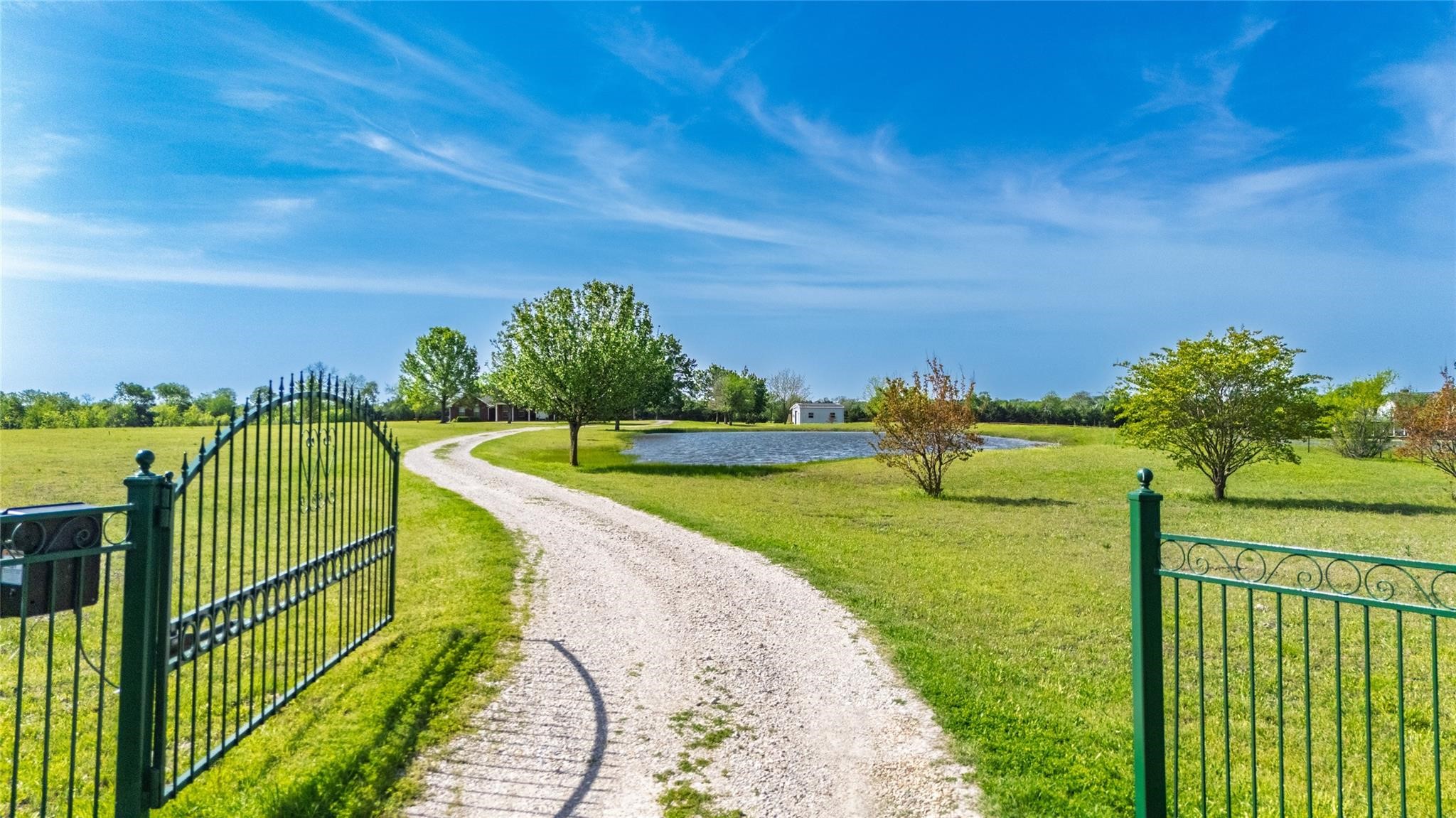 Rural property entrance with open metal gates, a curving gravel drive, and a pond beyond the lawn.
