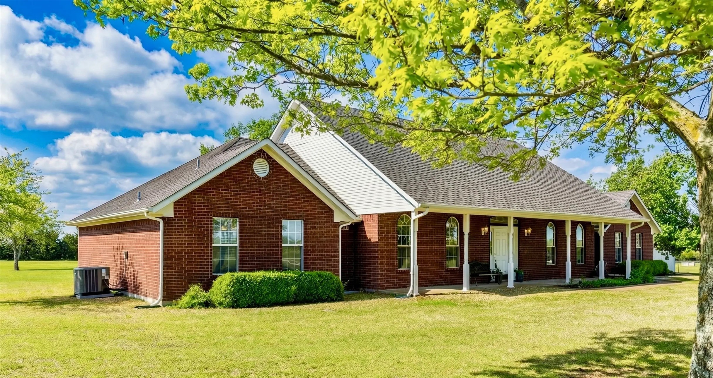 Red brick home with a long covered porch and broad lawn beneath a leafy tree.