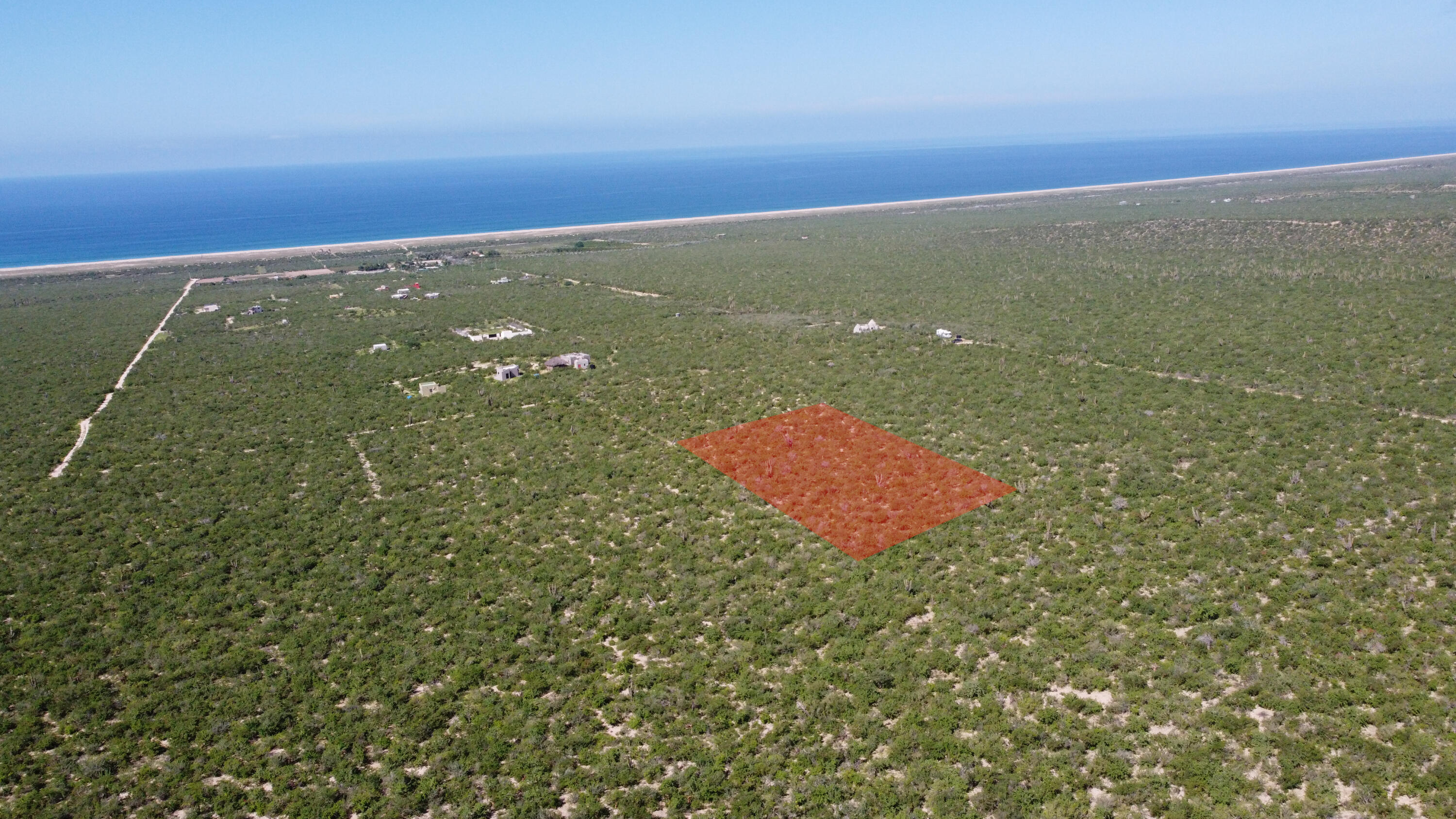 Aerial view of a red-outlined vacant lot amid coastal scrub, with the beach and ocean beyond.
