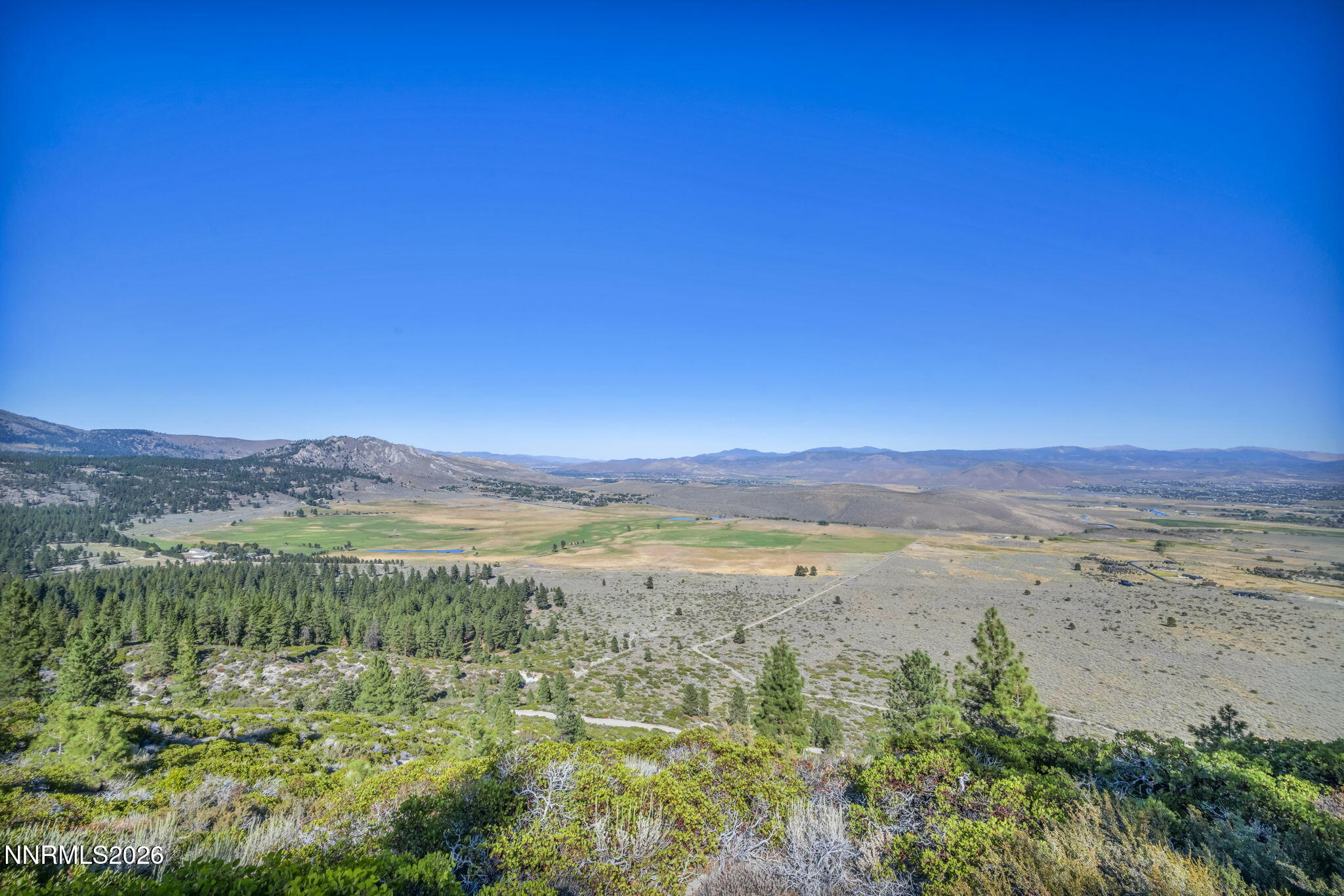 Panoramic valley view with open fields, pine-covered foothills, and distant mountain ranges.