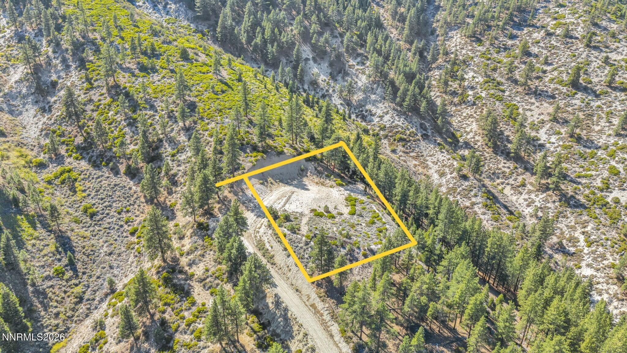 Aerial view of a vacant hillside parcel outlined in yellow, with a graded clearing, dirt track, and scattered pines.
