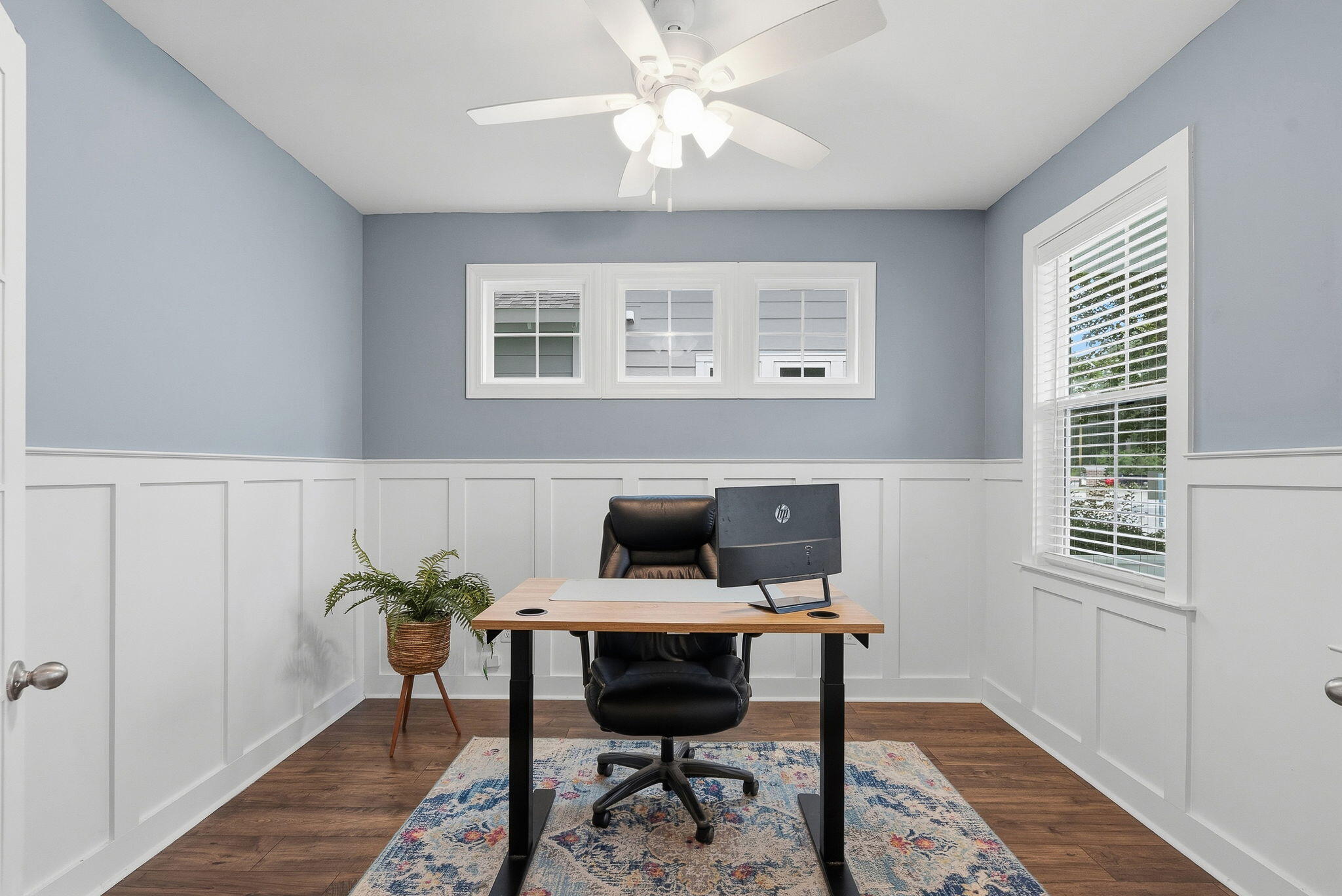 Furnished home office with a wooden desk, black chair, paneled walls, and windows.