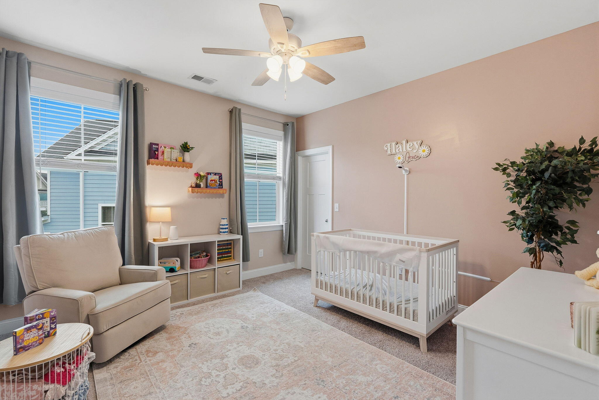 Nursery with a white crib, blush-pink wall, beige glider, and ceiling fan.