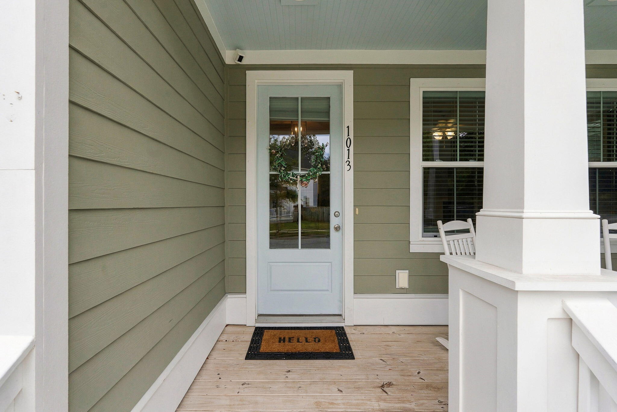 Covered front porch with a pale blue glass-paneled door, sage siding, white columns, and a welcome mat.