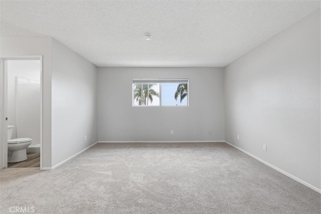 Empty carpeted bedroom with a centered window and an adjoining bathroom doorway.