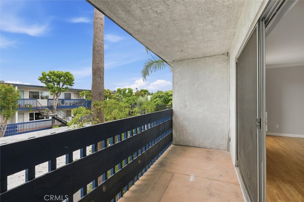 Covered apartment balcony overlooking landscaped grounds and neighboring buildings, with sliding glass doors.
