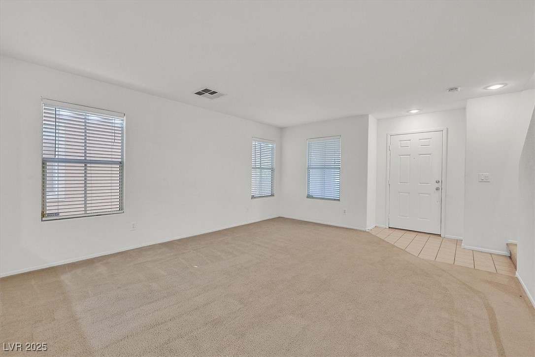 Empty living room with beige carpet, three blinded windows, and a tiled entry by the front door.