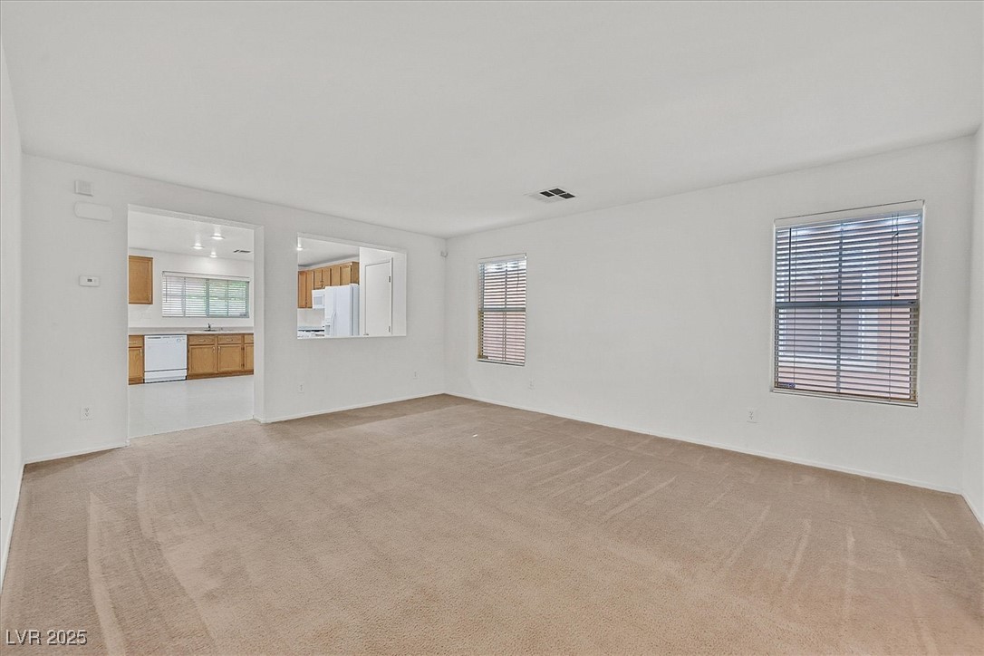 Unfurnished living room with beige carpet and two blind-covered windows, opening to a kitchen.