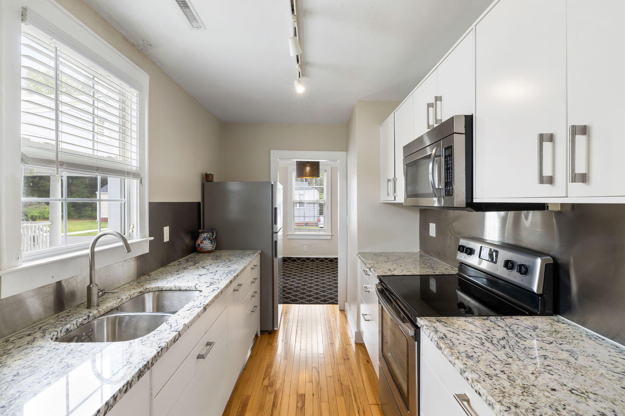 Galley kitchen with white cabinets, granite counters, a double sink, stainless steel appliances, and hardwood floors.