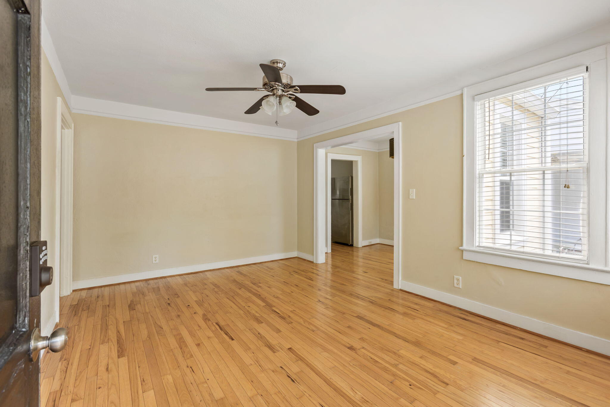 Empty room with hardwood floors, pale yellow walls, ceiling fan, and a large window with blinds.