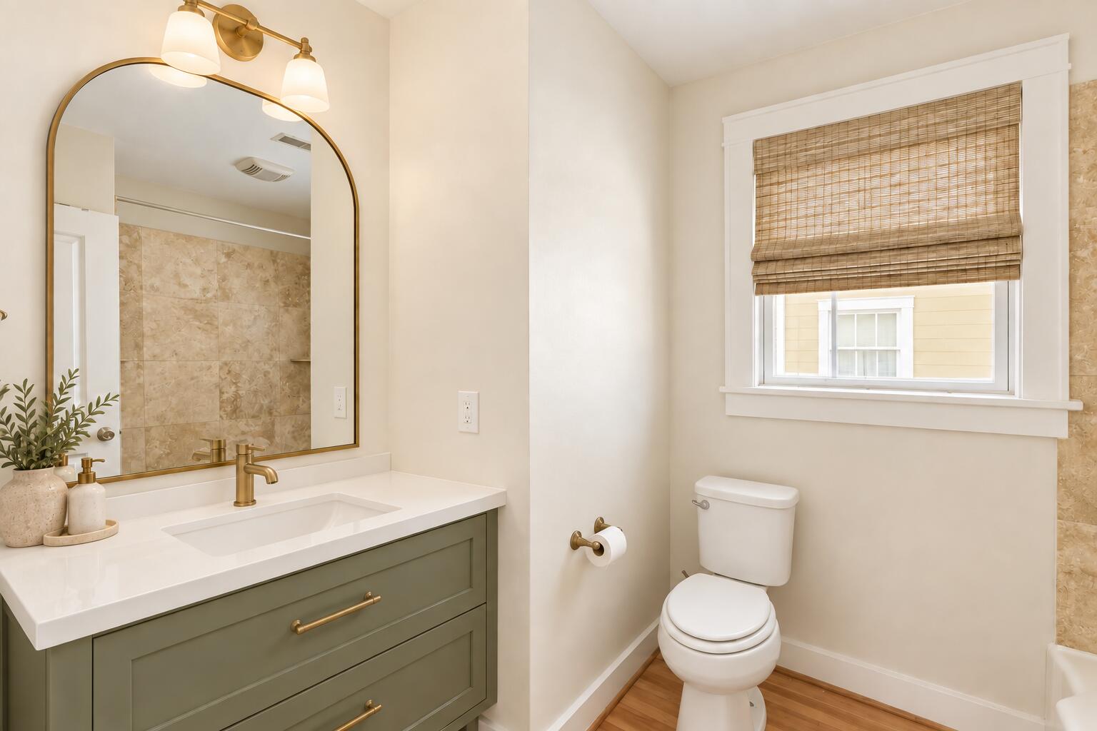 Bathroom with a green vanity, arched brass-framed mirror, and woven shade over the window.