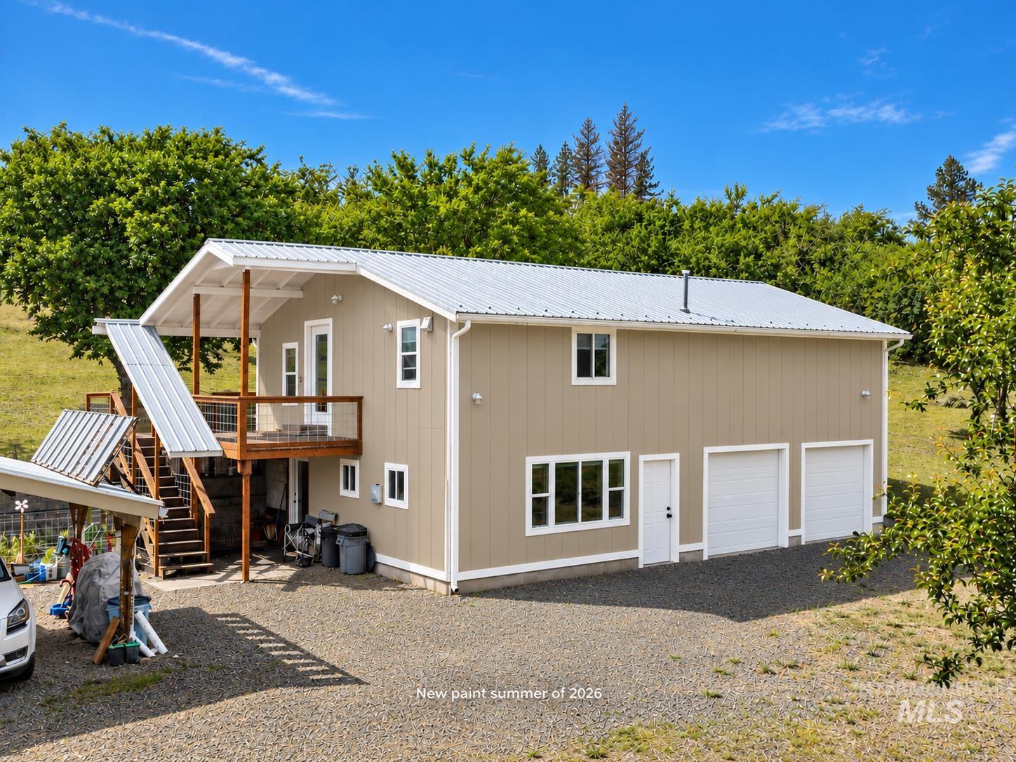 Beige rural home with a white metal roof, two garage doors, and an elevated covered deck.