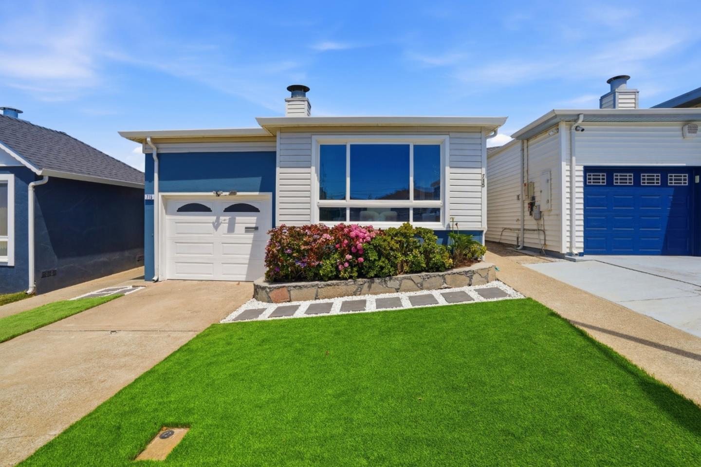 Single-story blue-and-white home with a large front window, attached garage, and flower bed.