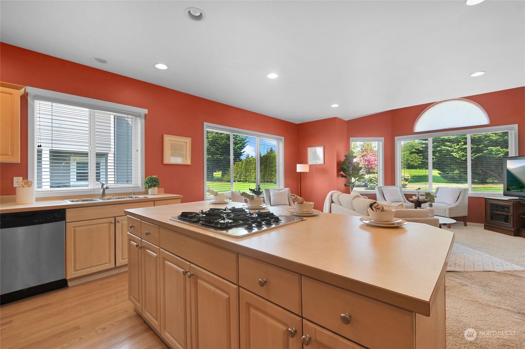 Kitchen with a large gas cooktop island, light wood cabinets, and adjoining living area with garden-facing windows.