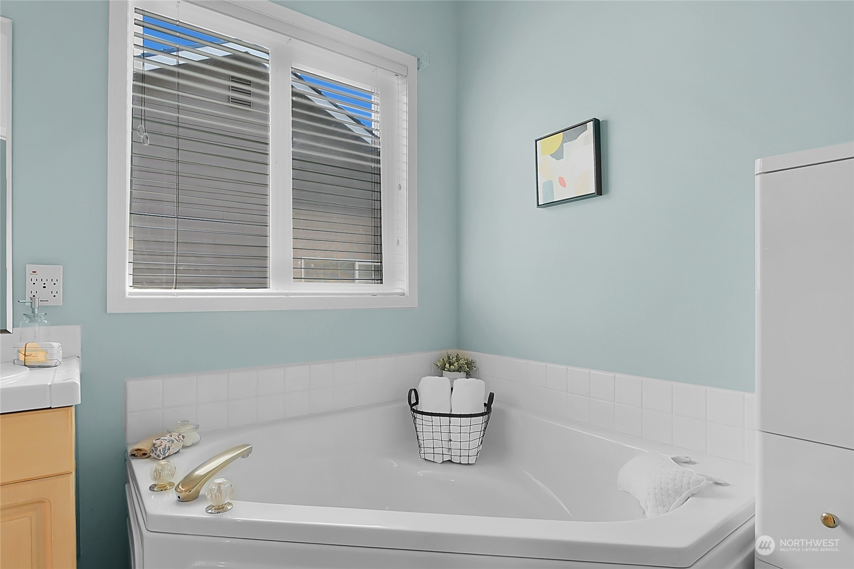 Bathroom with a large corner soaking tub, pale blue walls, and a window with blinds.