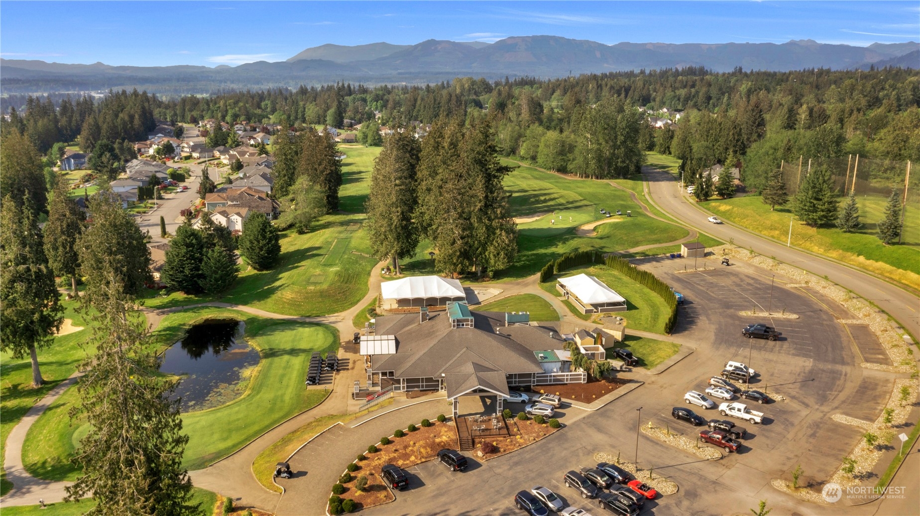Aerial view of a golf course clubhouse, greens, pond, and distant mountains.