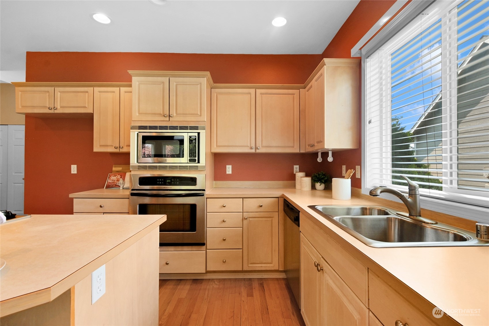Kitchen with light wood cabinets, stainless appliances, terracotta walls, and a sink beneath a large window.