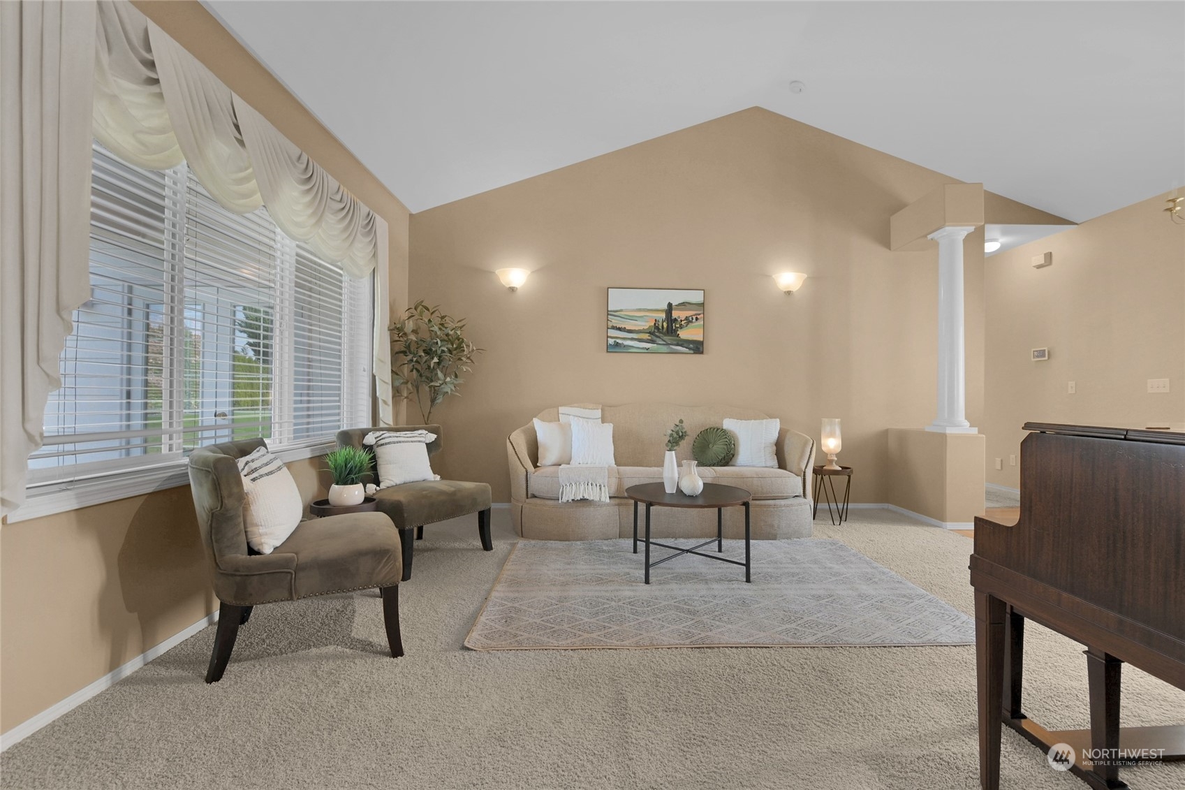Staged living room with vaulted ceiling, large windows, neutral seating, and an upright piano.