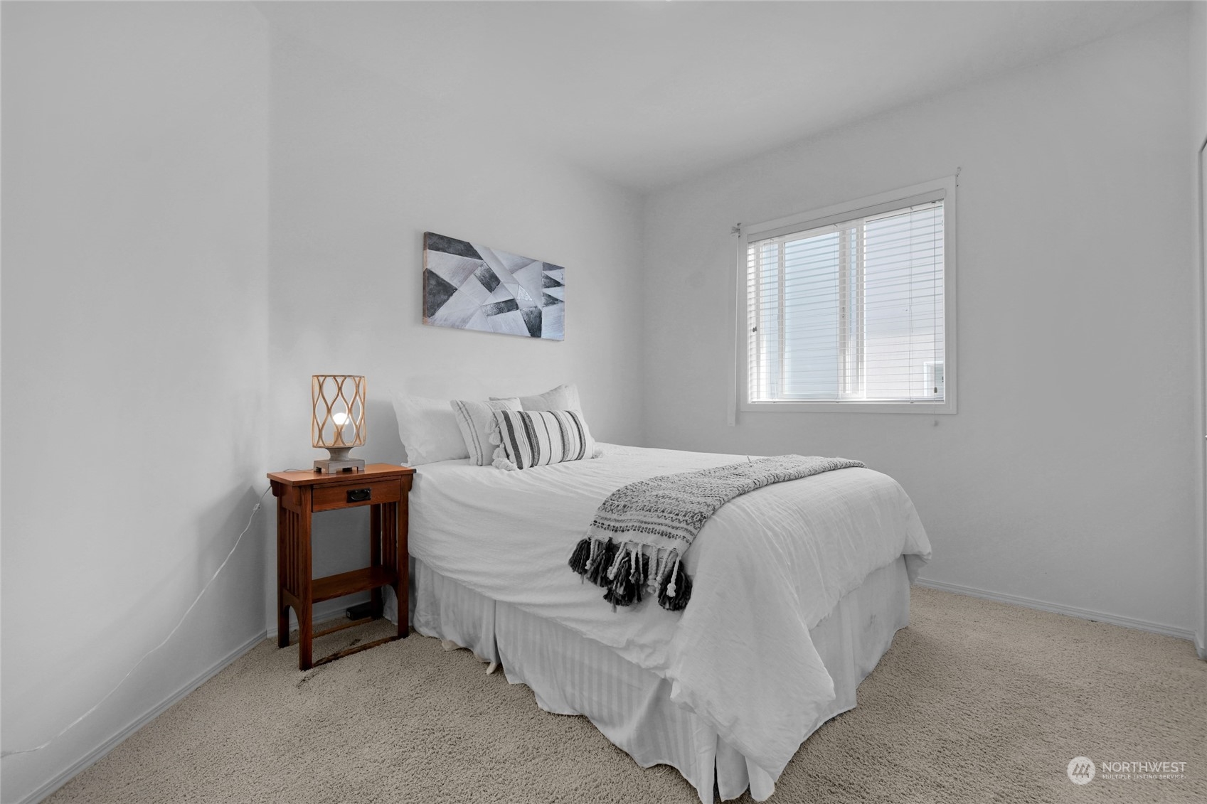 Staged bedroom with a white bed, patterned throw, wooden bedside table, and window with blinds.
