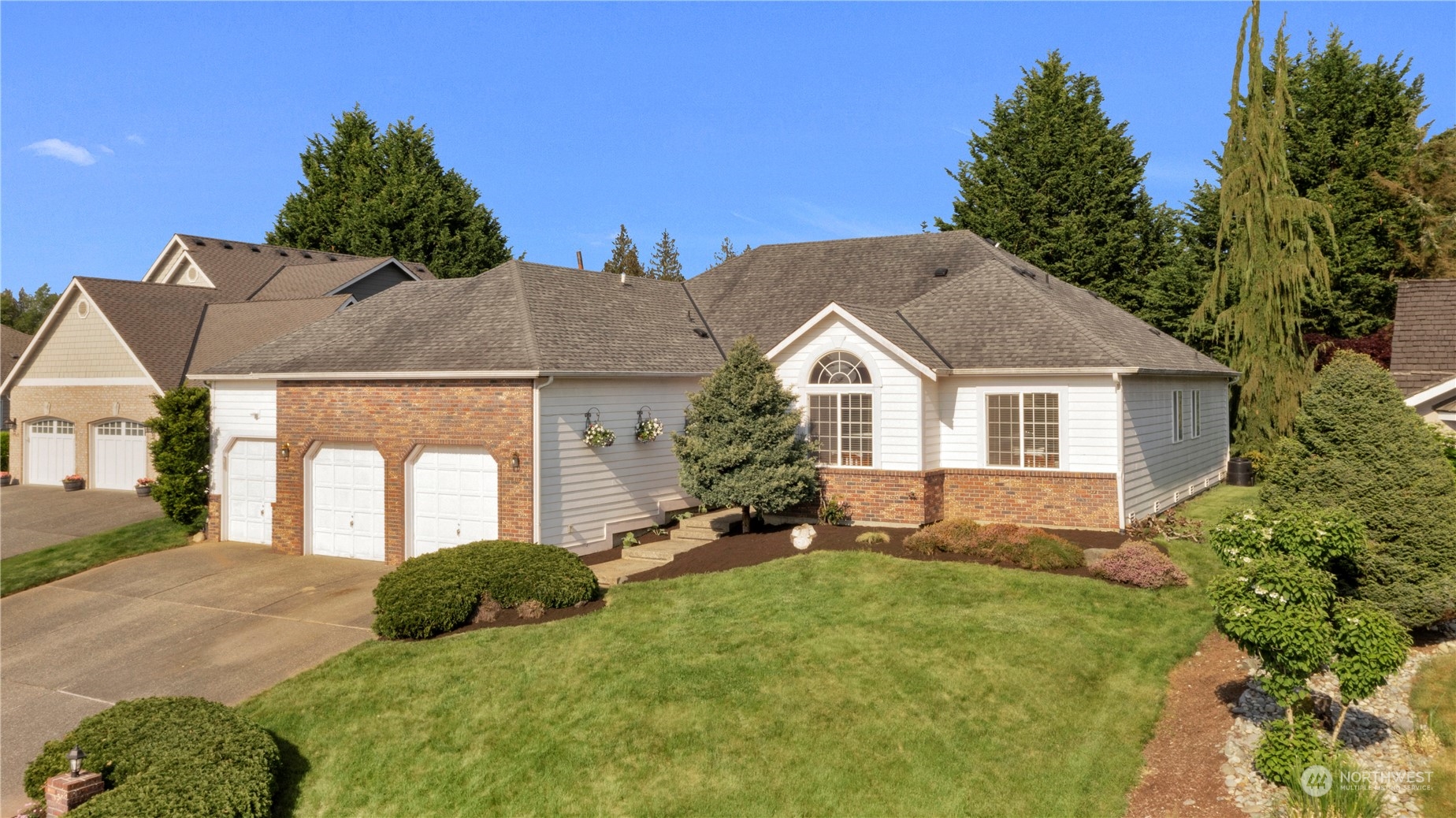 Home exterior with brick-accented three-car garage, landscaped lawn, and mature evergreens.